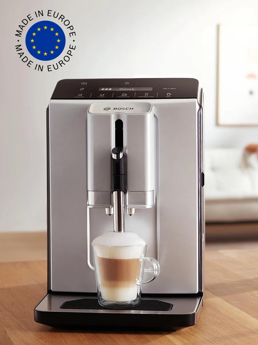 Aparat VeroCafe pe blatul de bucătărie, cu o ceașcă de cappuccino și steagul european în colțul de sus al imaginii.