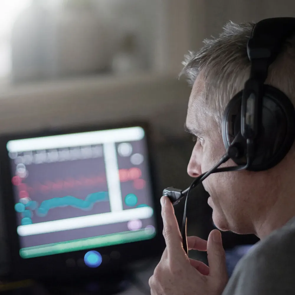 Un uomo dai capelli grigi, con indosso un headset dotato di microfono, osserva lo schermo di un computer che mostra grafici finanziari e dati sfocati, in una stanza buia.