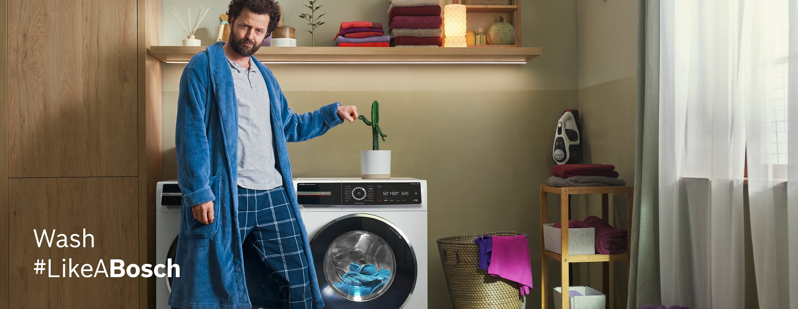 Man in blue robe standing next to washing machine in home interior with shelves and decor.