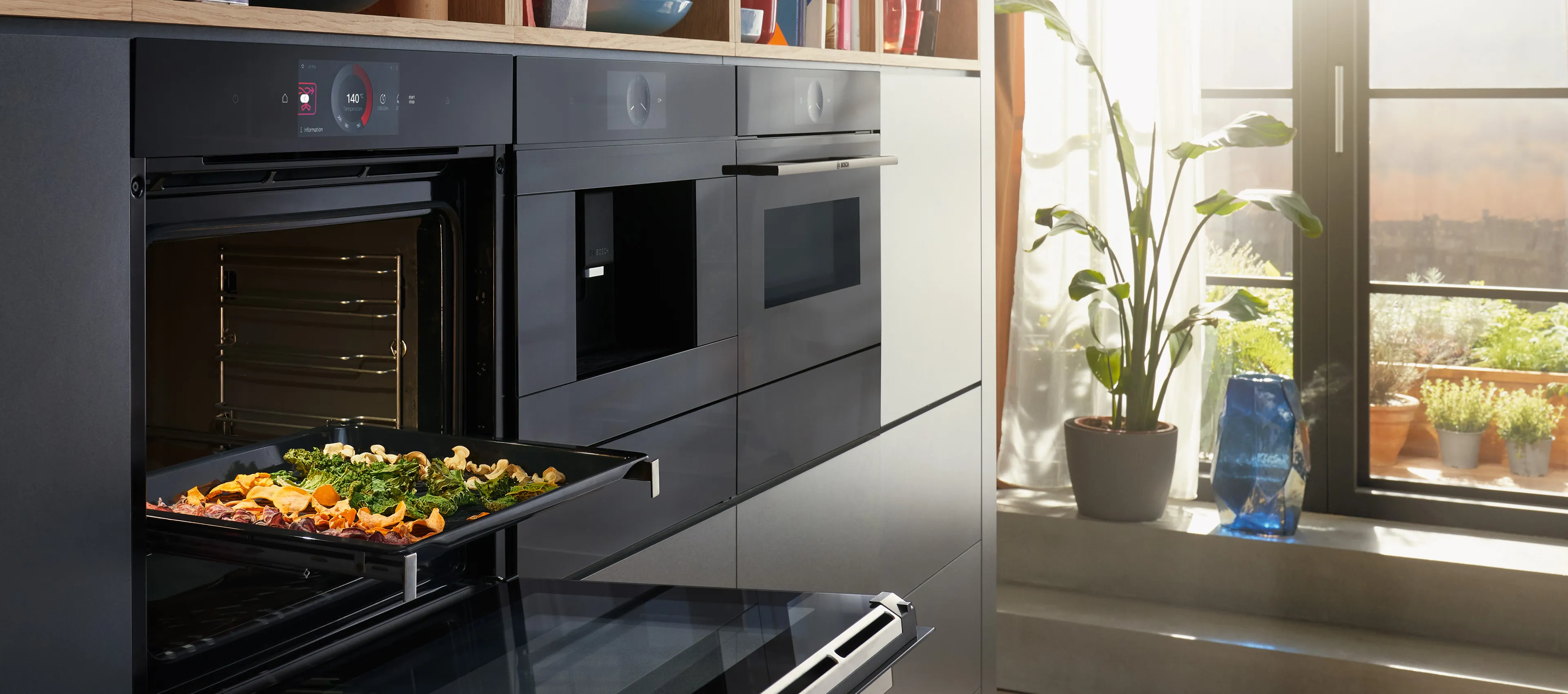 Moderne, dunkle Einbauküche mit einer Gerätezeile aus Bosch Serie 8 Einbauten, darunter ein geöffneter Backofen mit einem Blech Ofengemüse.