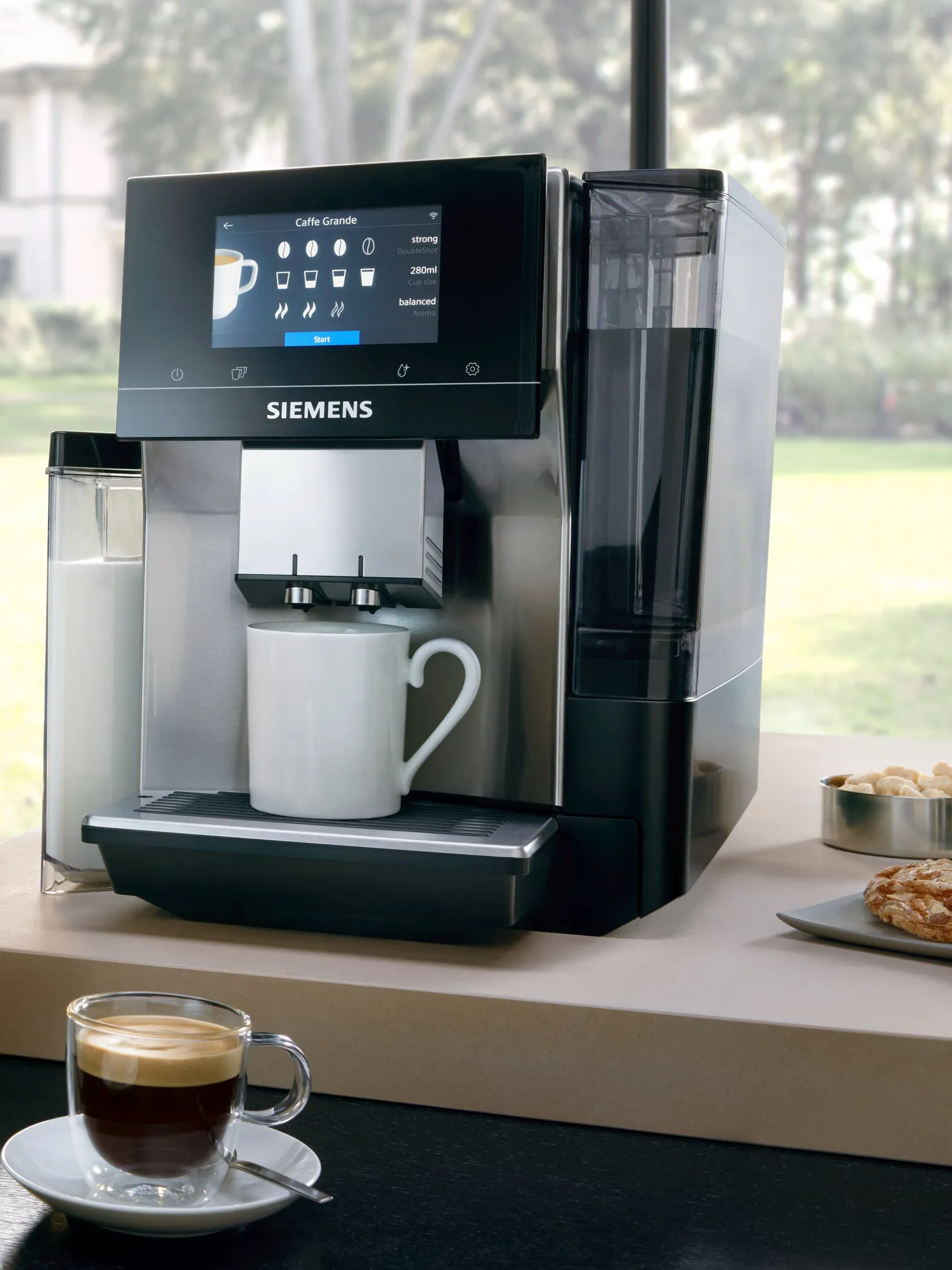 Coffee machine with touchscreen display, milk frother, and a cup of coffee on a table.