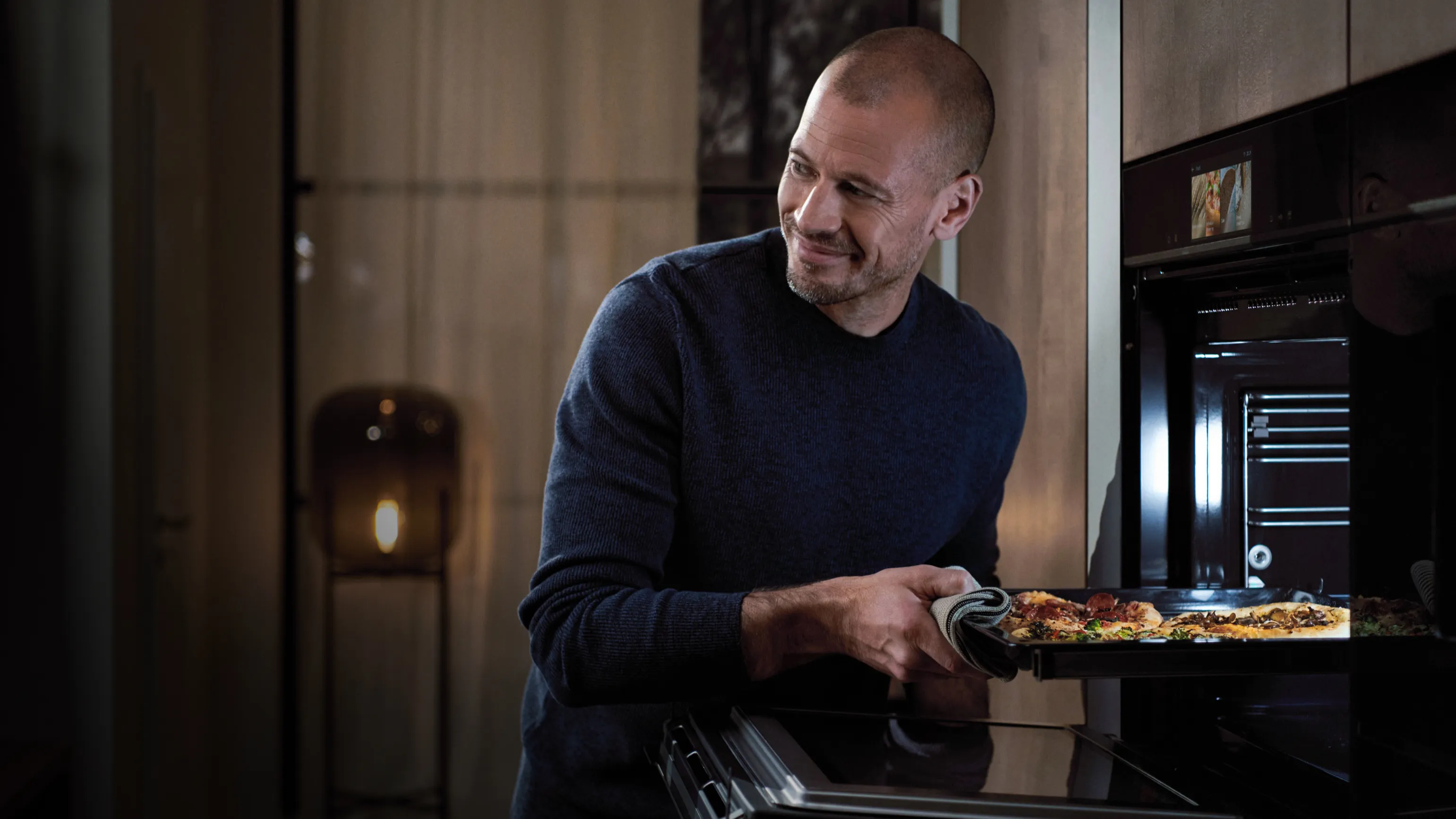 Uomo in maglione scuro prendendo pizza appena cotta dal forno in ambiente accogliente cucina.