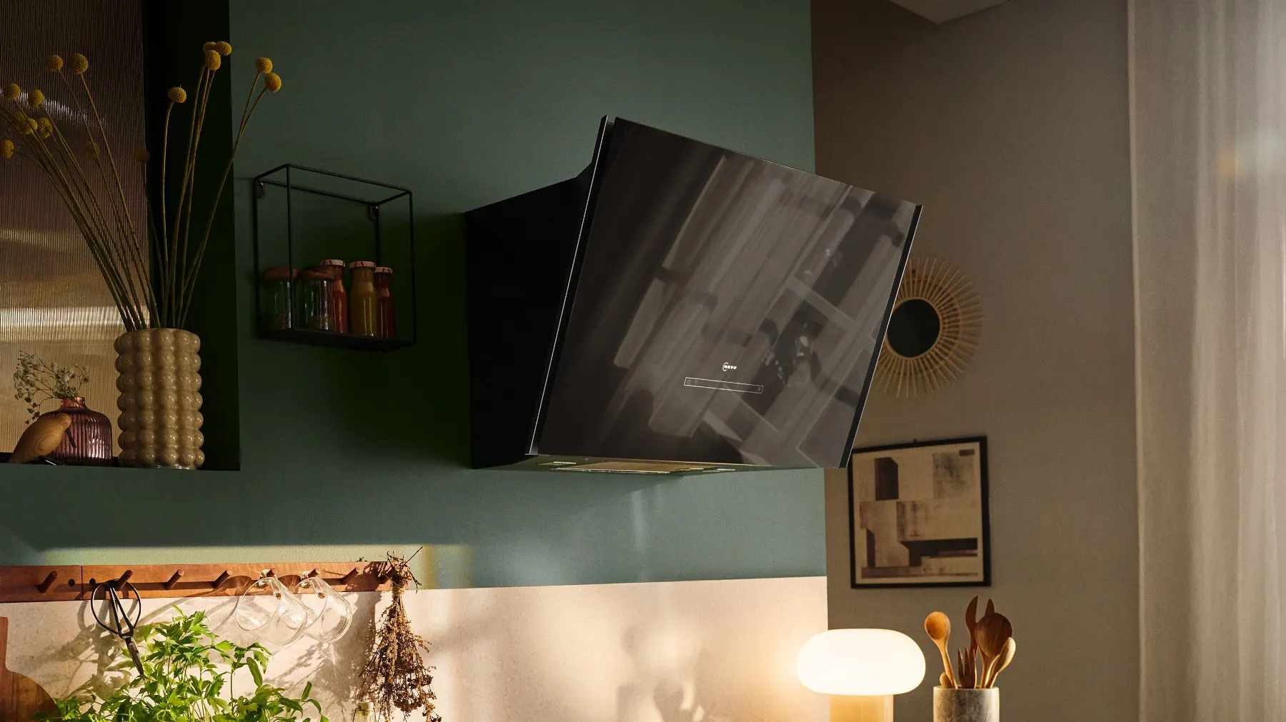 Moderne Küche mit schräg montierter Glas-Dunstabzugshaube über dem Kochfeld; auf der Arbeitsplatte frische Kräuter, Kochutensilien und eine Lampe, offene Regale mit Gläsern und Deko vor einer grünen Wand und ein gerahmtes Bild an der Wand.