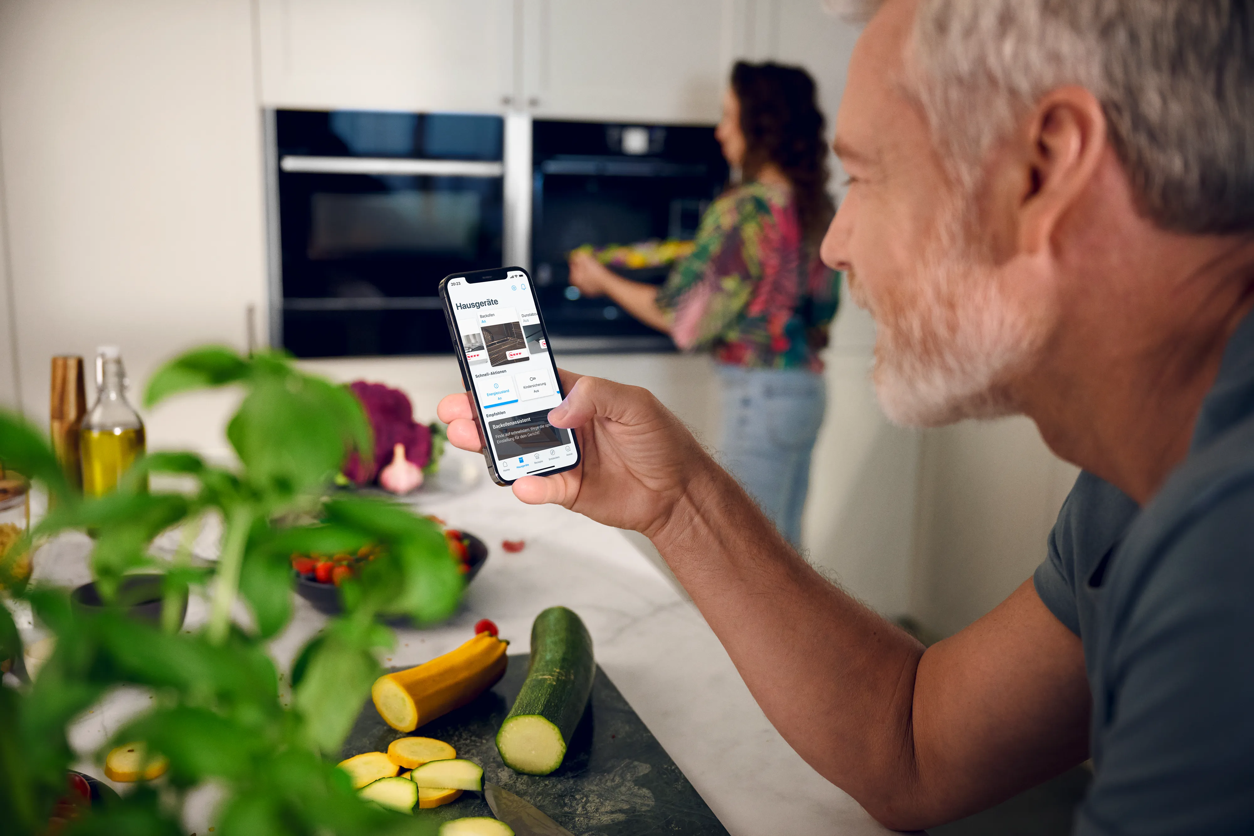Човек разглежда приложението Home Connect на телефона си в модерна бяла кухня
