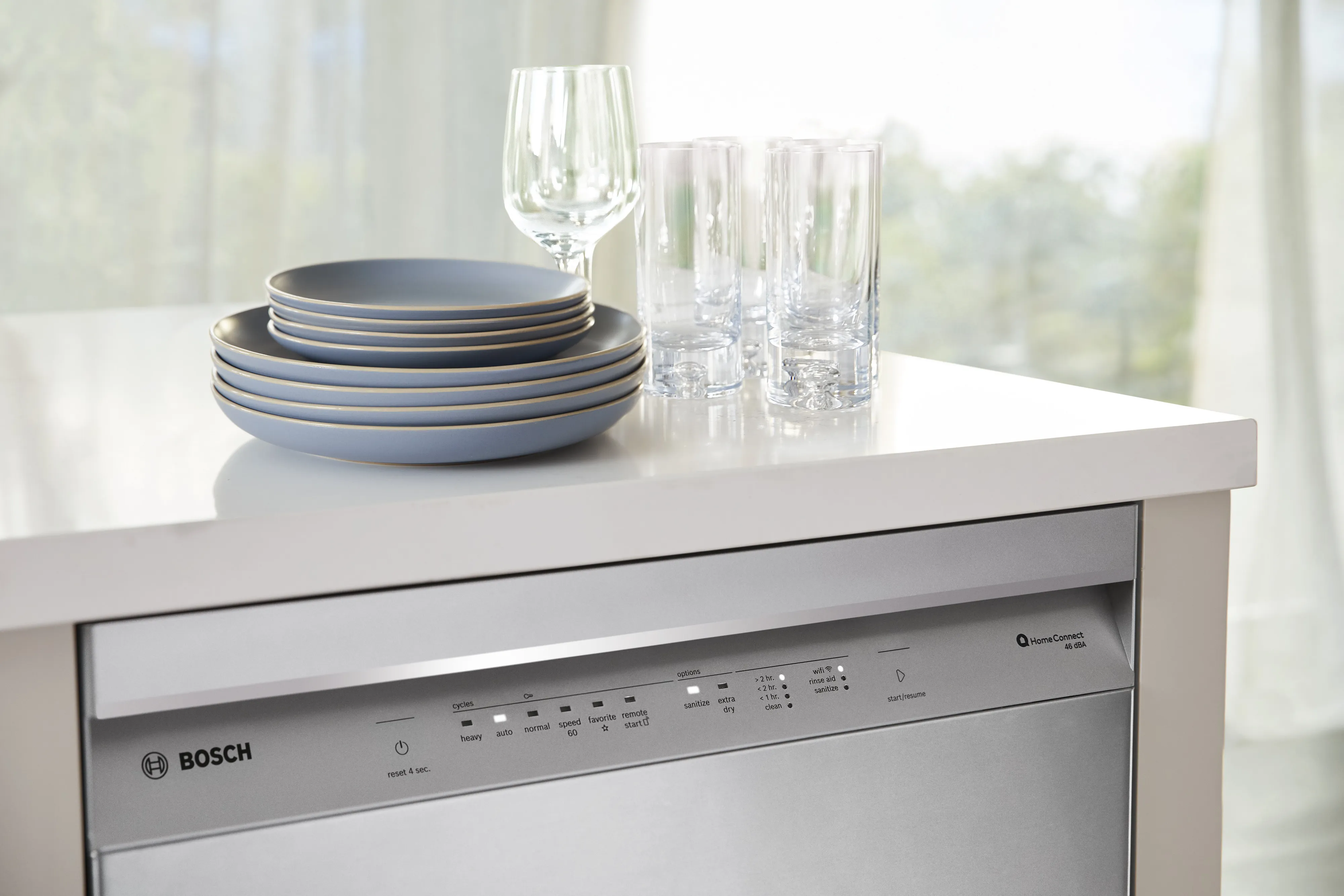 Bosch dishwasher with control panel, dishes, wine glass, and tumblers on a white countertop.