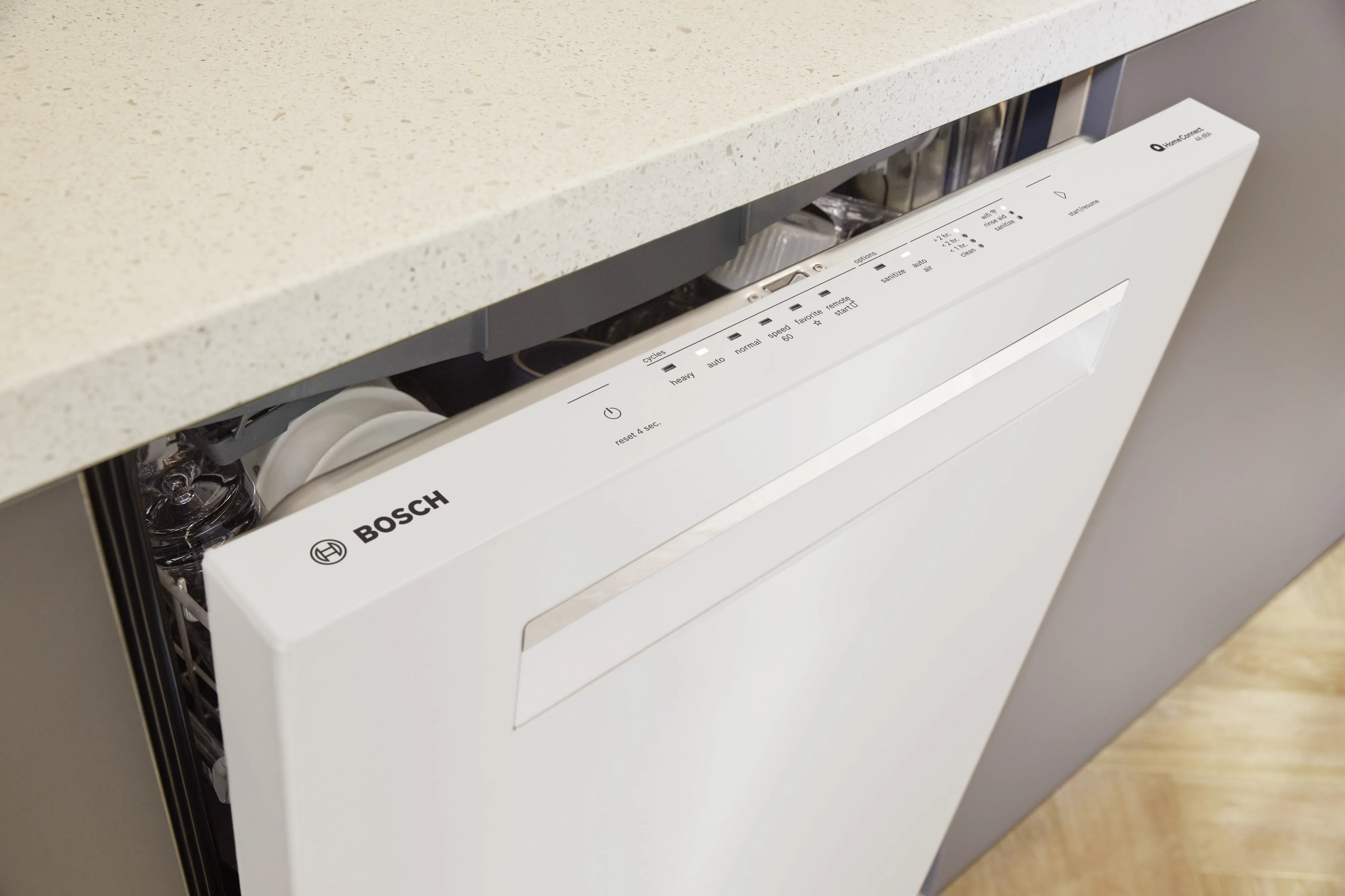Bosch white dishwasher partially open under a gray countertop, showing control panel and dishes.