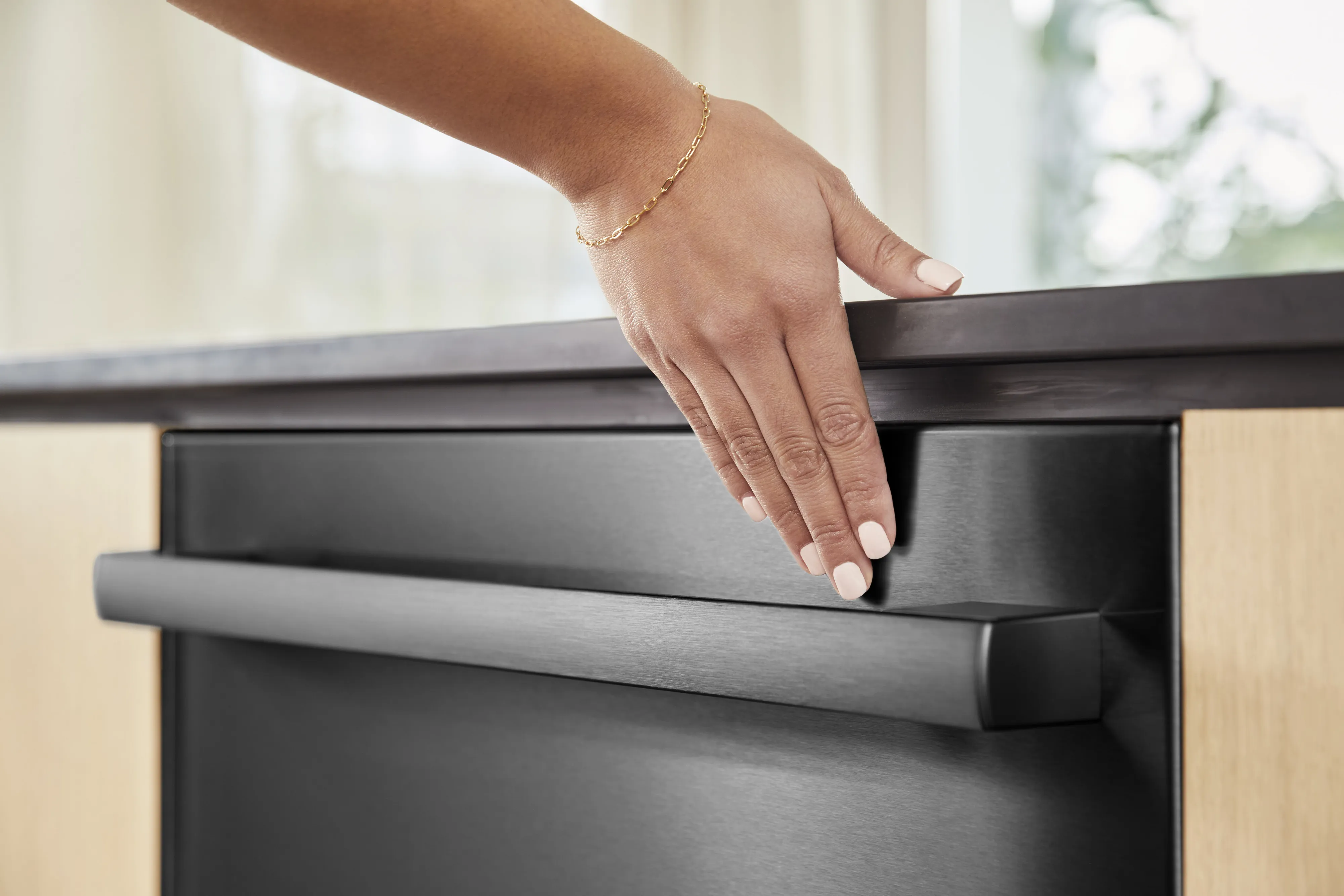Hand reaching to open the handle of a modern Bosch black dishwasher in a kitchen setting.