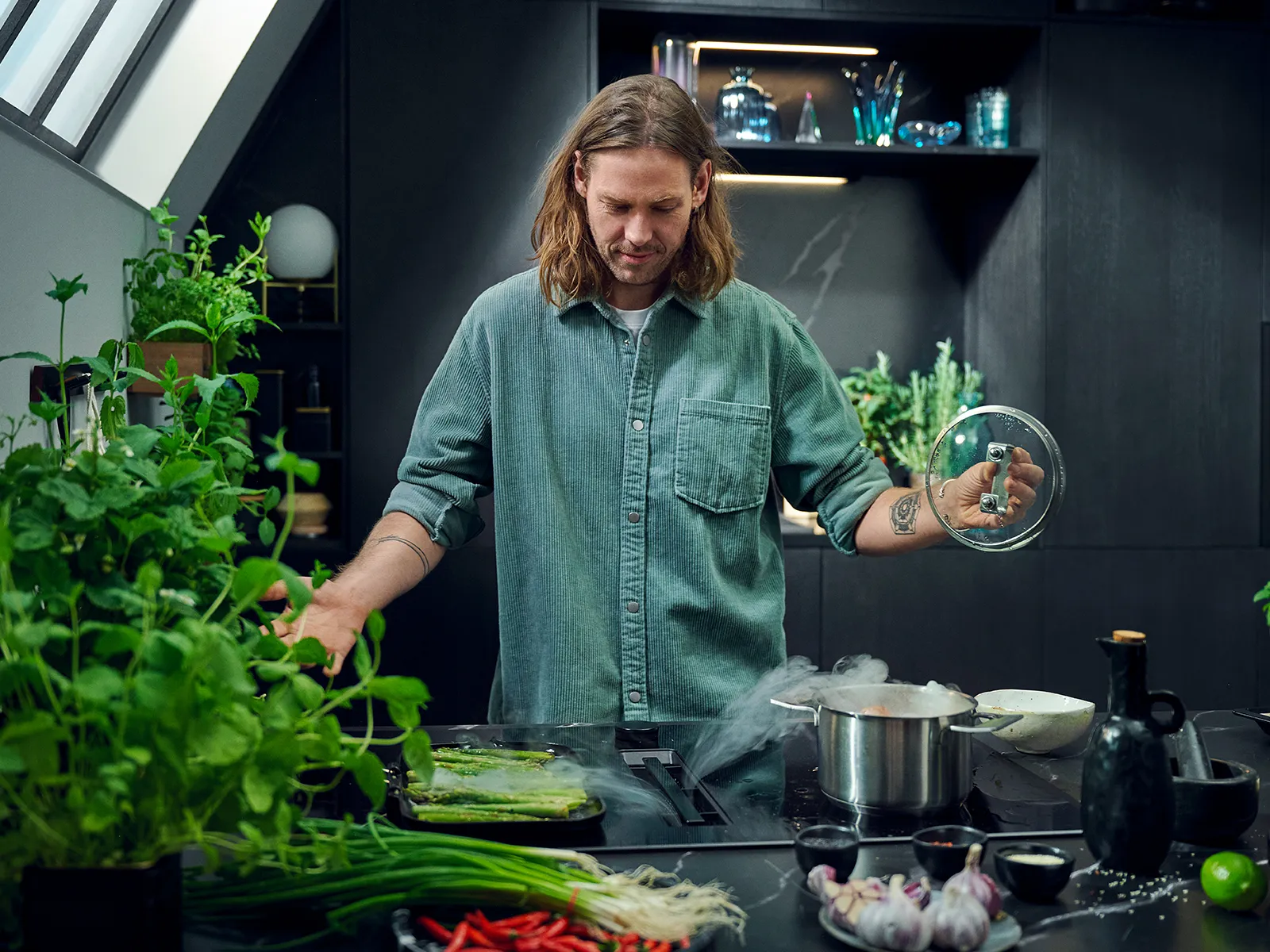 Man cooking in kitchen surrounded by fresh vegetables and plants.