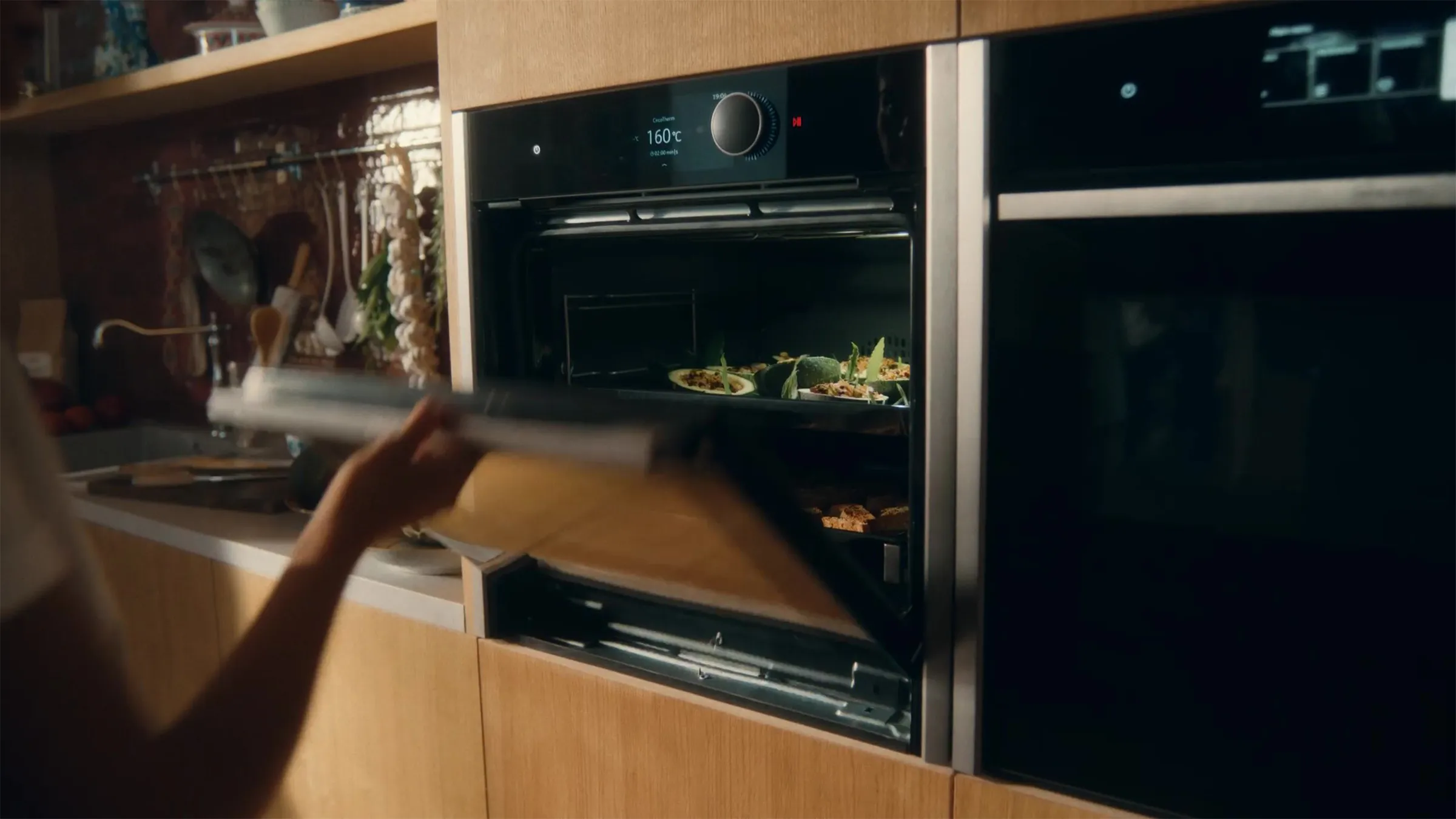 A person's arm and hand are visible as they open the door of a sleek, black built-in oven. Inside the oven, trays hold small, round food items, possibly stuffed vegetables or pastries. The oven has a digital display and is integrated into light wooden cabinetry with a red tiled backsplash and hanging kitchen utensils in the background. Another matching oven is visible to the right.