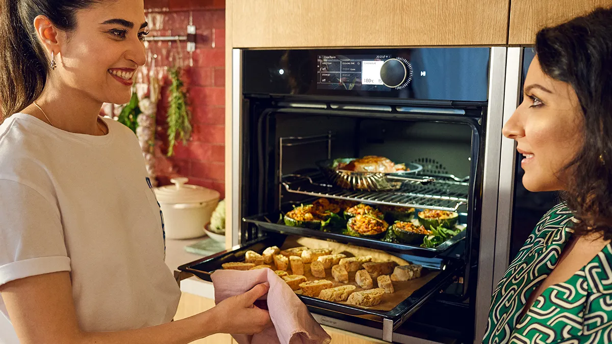 Two women are smiling and interacting in a modern kitchen. The woman on the left, with dark hair pulled back and wearing a white t-shirt, holds a baking tray of biscotti or similar baked goods, partially pulled out from a sleek, black built-in oven. The woman on the right, with dark, curly hair and wearing a green patterned top, looks towards the oven with a pleased expression.