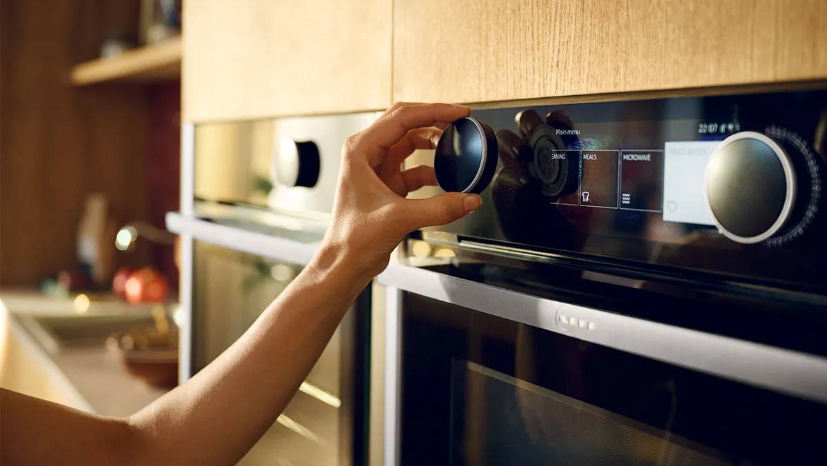 A close-up shot shows a hand adjusting a black rotary knob on the sleek, black control panel of a built-in oven. The digital display on the oven shows a "Main menu" with options like "DRYING", "MEALS", and "MICROWAVE", along with a clock displaying "22:07". The oven is integrated into light wooden cabinetry, and another matching oven is partially visible to the left. The background is softly blurred, showing hints of a modern kitchen interior.