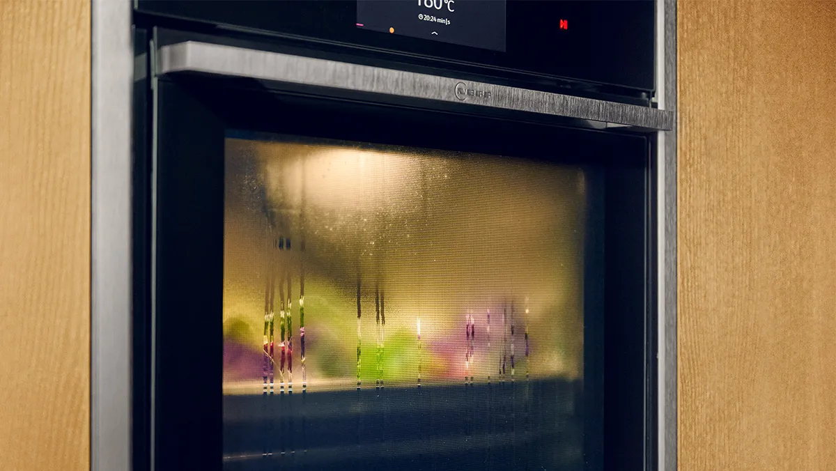 A close-up of a sleek, black built-in oven with a glass door showing condensation, indicating something is cooking inside. The digital display above the door shows "180°C" and a timer counting down from "20:24 min". The oven has a textured handle and is integrated into light wooden cabinetry.