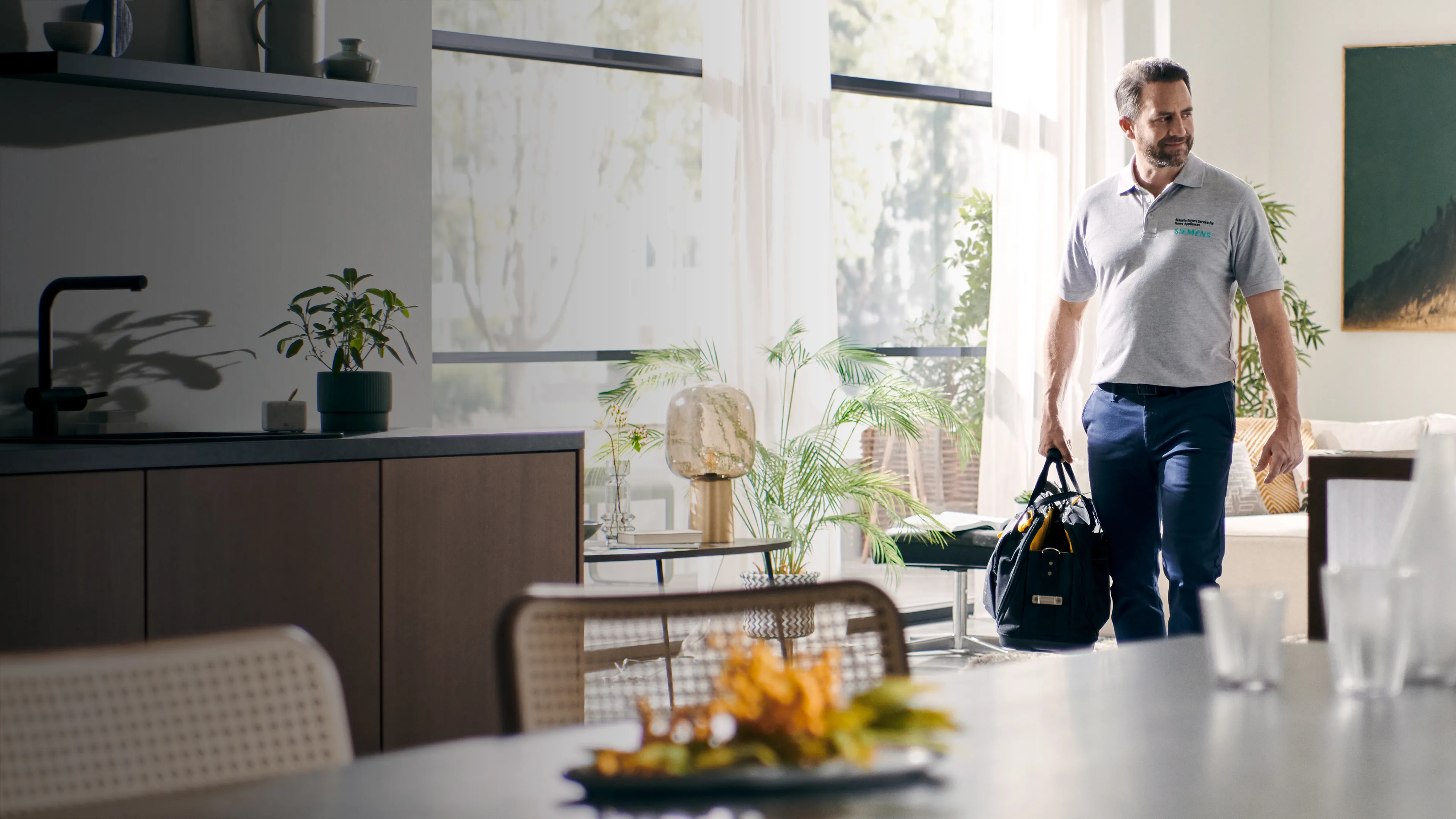 La imagen muestra a un hombre en una camisa de polo gris llevando una mochila, de pie en un moderno y bien iluminado salón con plantas y muebles.