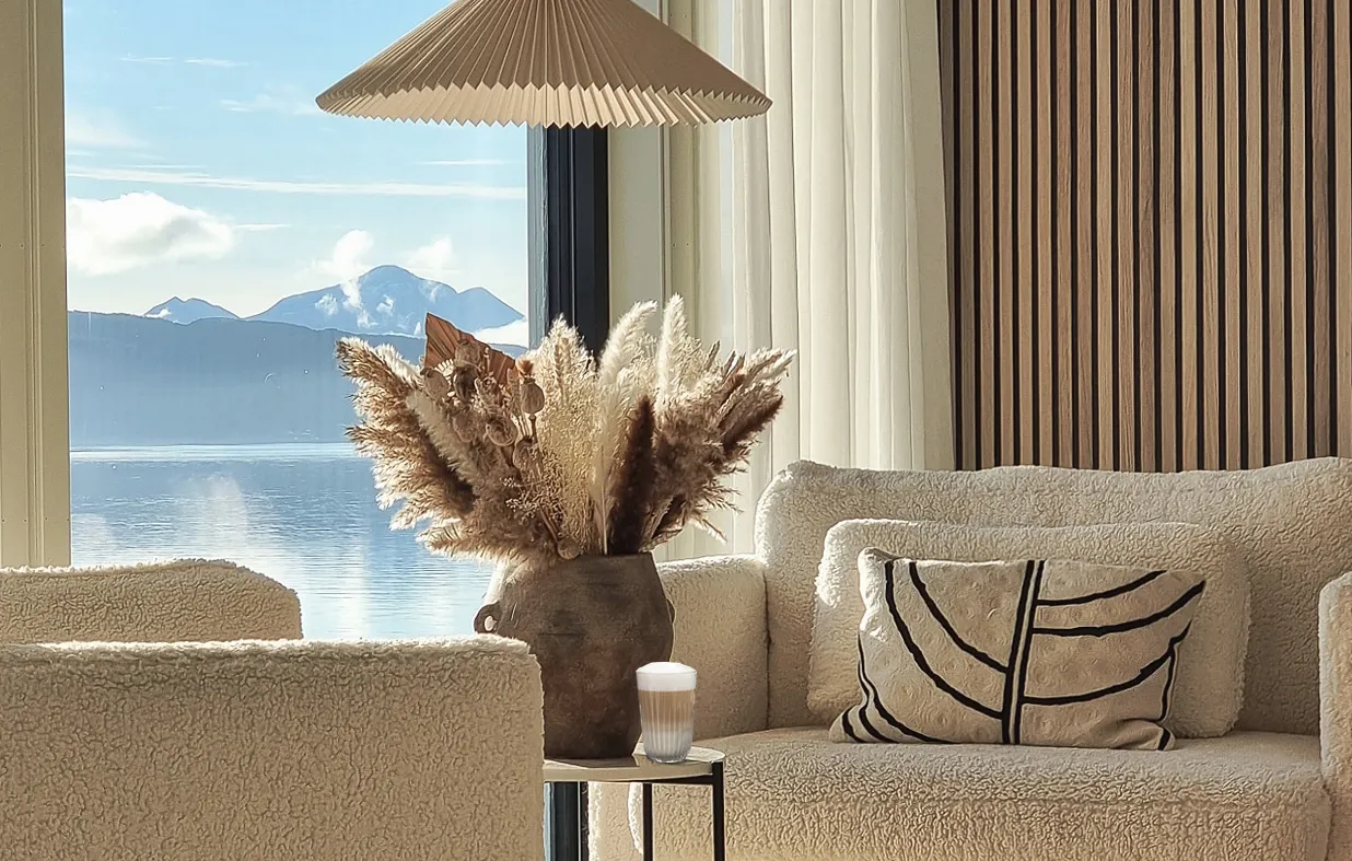 Hyggelig stue med bløde lænestole, en dekorativ vase med pampasgræs, og en naturskønt udsigt til bjerge og vand gennem et stort vindue.