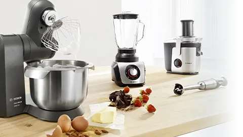 Kitchen appliances including mixer, blender, and food processor on wooden table with ingredients.