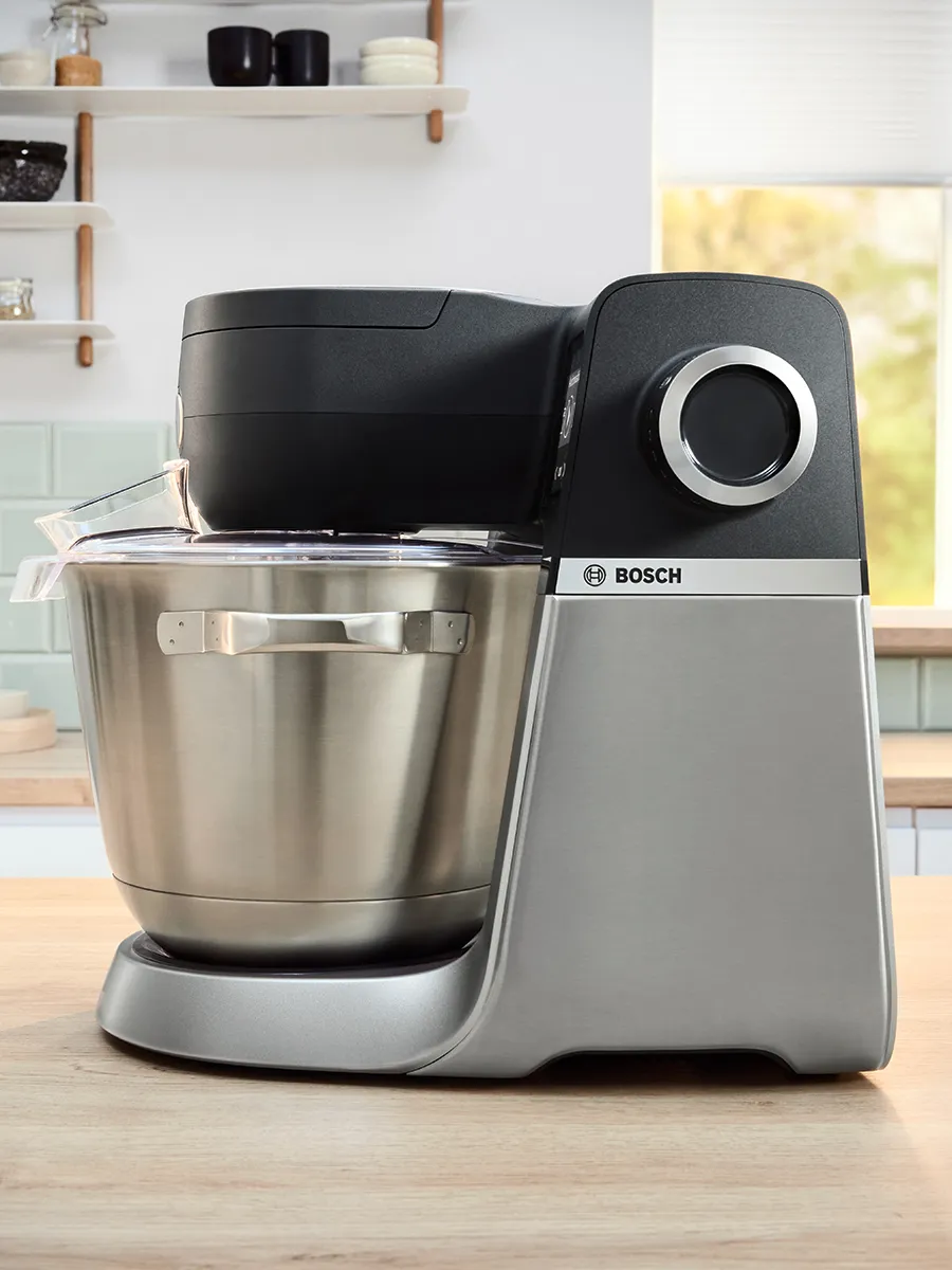 Un moderno mixer da cucina Bosch con ciotola, fruste e accessori in acciaio inox, posizionato su un piano di lavoro in legno in una luminosa cucina.