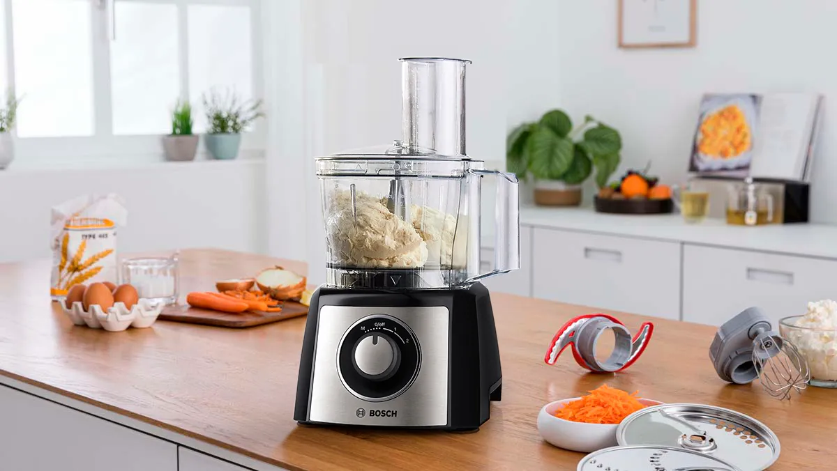 Procesador de alimentos Bosch sobre una encimera de cocina, rodeado de verduras, huevos, utensilios e ingredientes preparados.