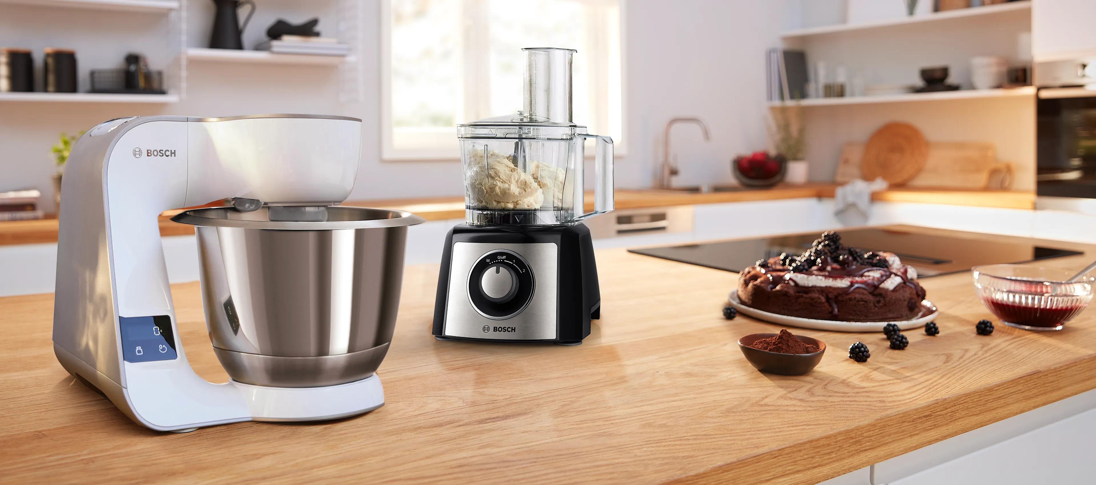Batidora de pedestal Bosch y procesador de alimentos Bosch sobre una encimera de madera al lado de un pastel.