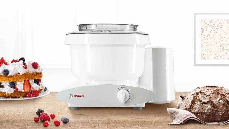 Bosch kitchen mixer in white on a wooden counter with cake and bread nearby.