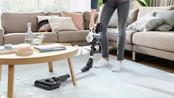 A woman uses a Bosch cordless vacuum cleaner to clean up white debris on a light-colored rug in a modern living room with a sofa and coffee table.