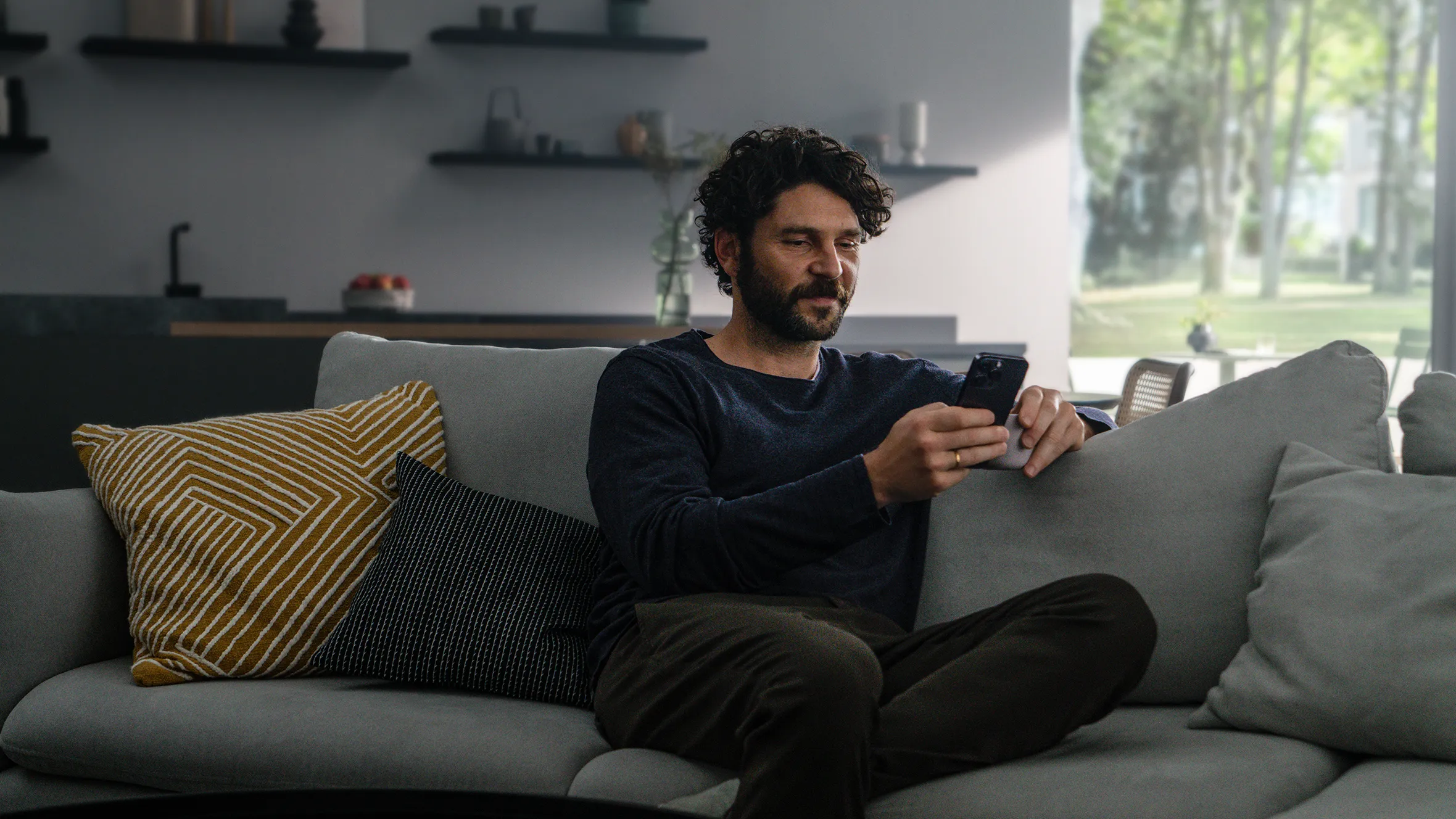 Akıllı telefon kullanarak kanepede oturan adam, yastıkla çevrili, modern oturma odasında pencere görünümü ile.