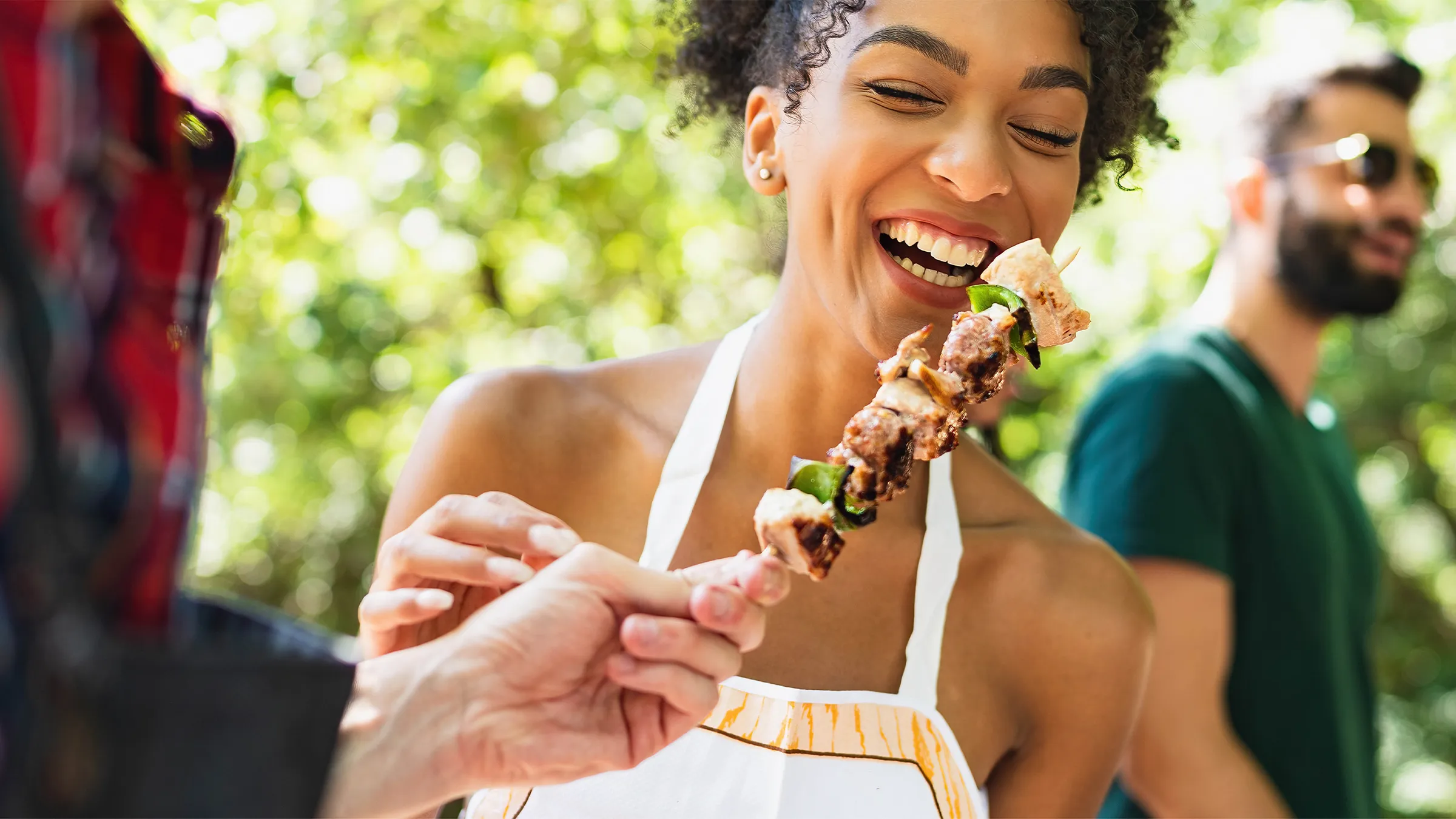 Smiling woman eating grilled food outdoors with friends in background.