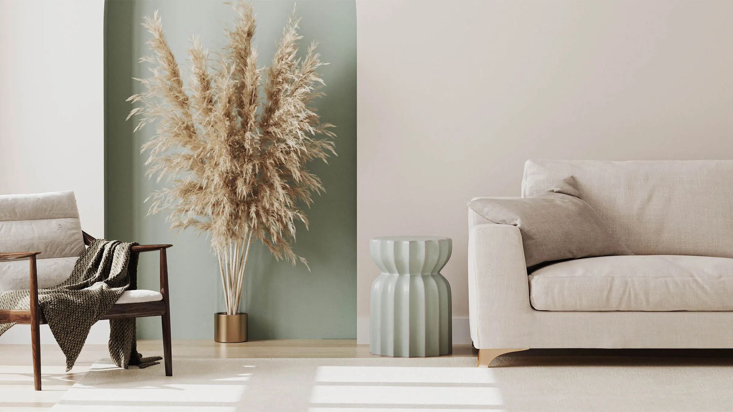A serene and stylish living room with neutral-toned furniture, including a light-colored sofa and a wooden armchair with a throw blanket draped over it. A large vase of fluffy pampas grass stands in a sage green wall nook, adding a natural, textural element to the calm, modern space.