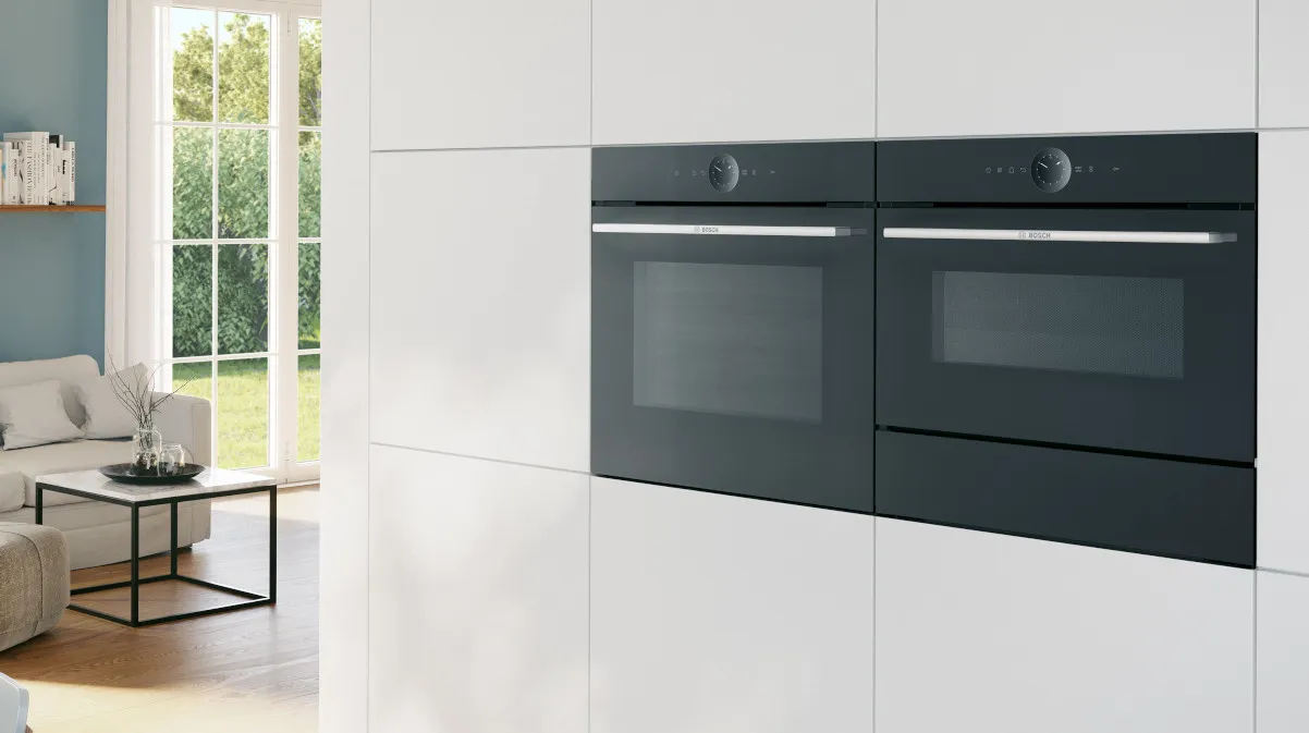 Moderne Küchenausstattung mit weißen Schränken, schwarzen Öfen und großem Fenster mit Blick auf die Außenlandschaft.