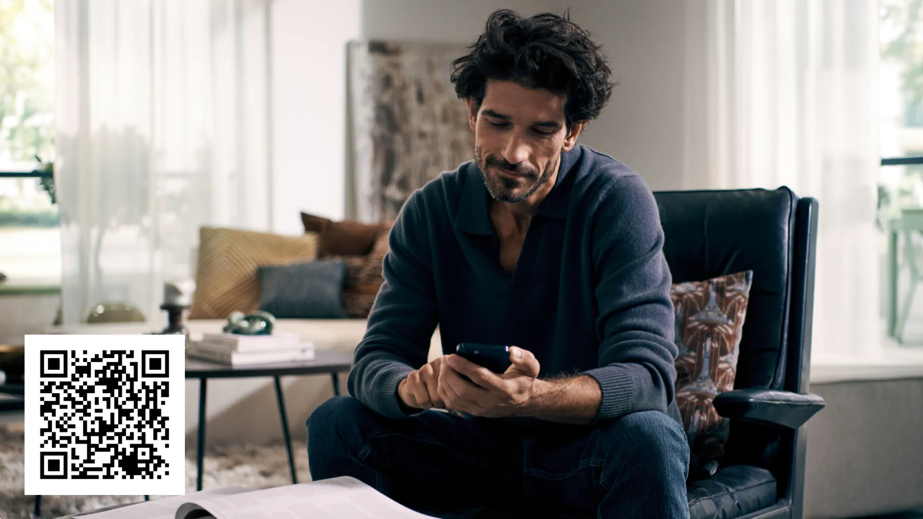 Homme assis dans un fauteuil utilisant un smartphone dans un salon lumineux, avec un code QR affiché en bas à gauche.