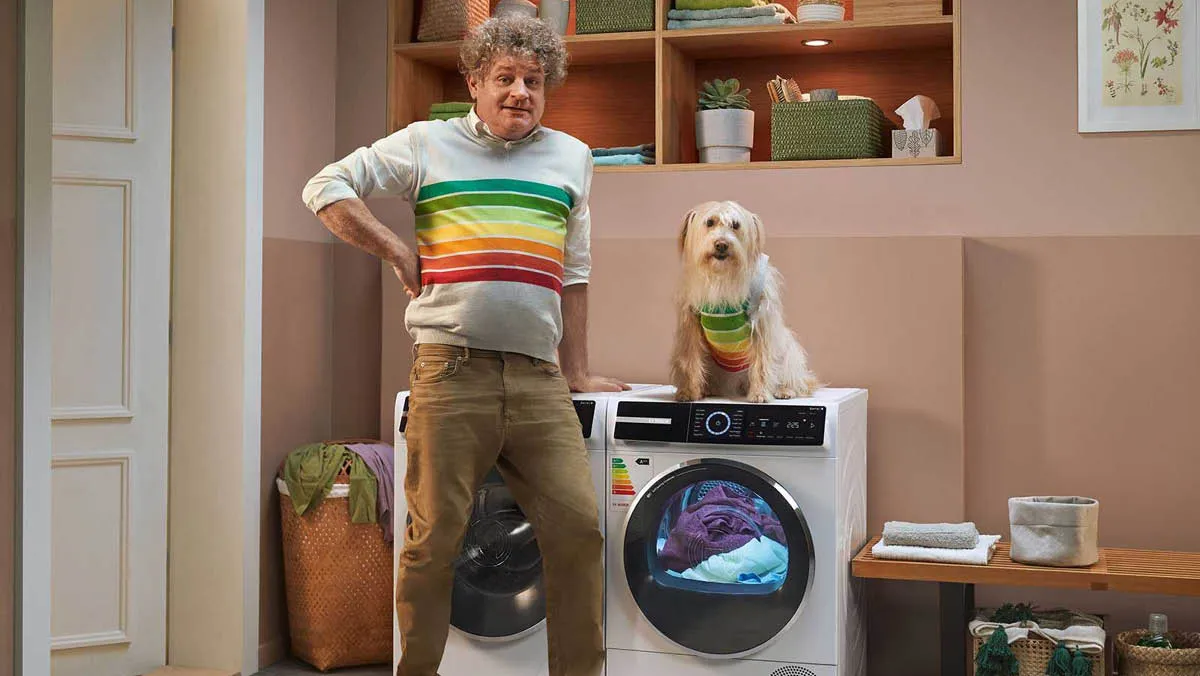 Mann im bunten Pullover steht mit Hund neben Waschmaschine im Hausinneren.