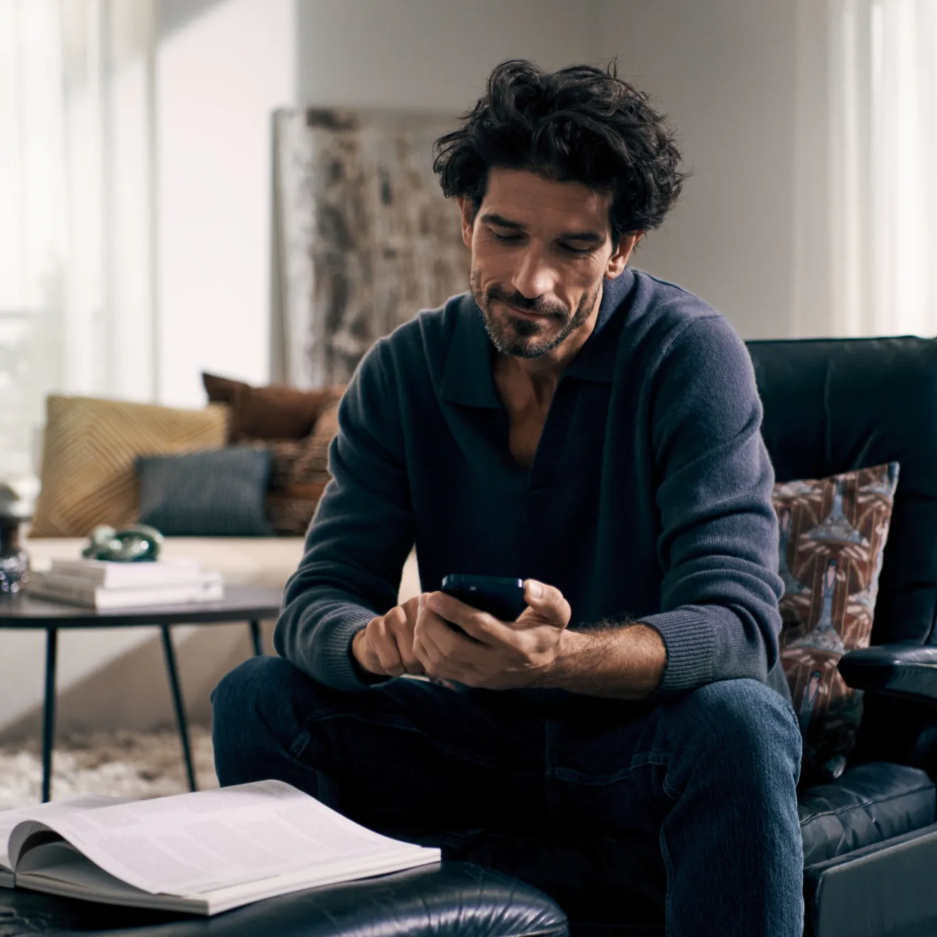 Mann mit lockigen Haar sitzen auf Couch, mit Smartphone, umgeben von Büchern und Dekor.
