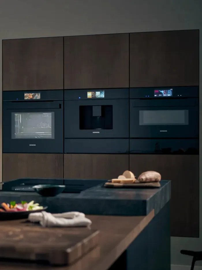 A kitchen scene featuring built-in Siemens iQ700 studioLine appliances, including an oven, a coffee machine, and a microwave, arranged horizontally within dark cabinetry