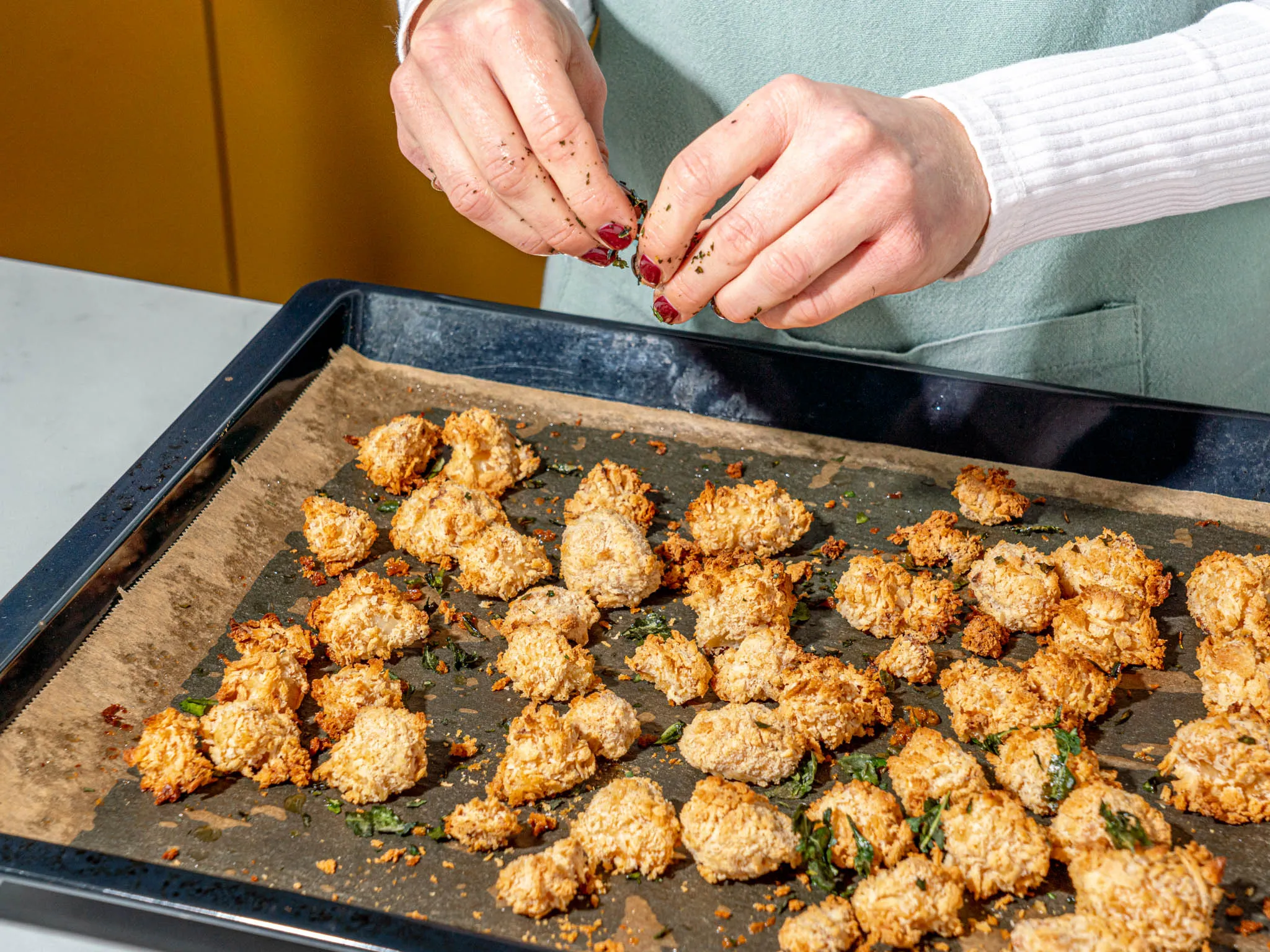Hands sprinkling herbs over freshly baked, golden-brown coconut macaroons on a parchment-lined baking tray.