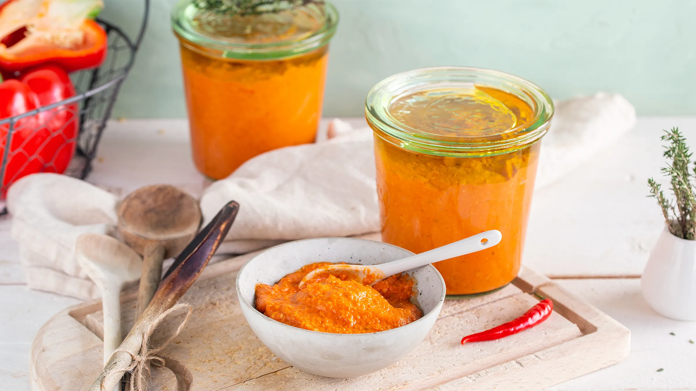 Pesto ilə dolu iki şüşə bankanın yanında taxta qaşıqlar və bir qırmızı çili bibəri olan kəsmə taxtanın üzərində keramik qabın içində qızardılmış bibər pesto.