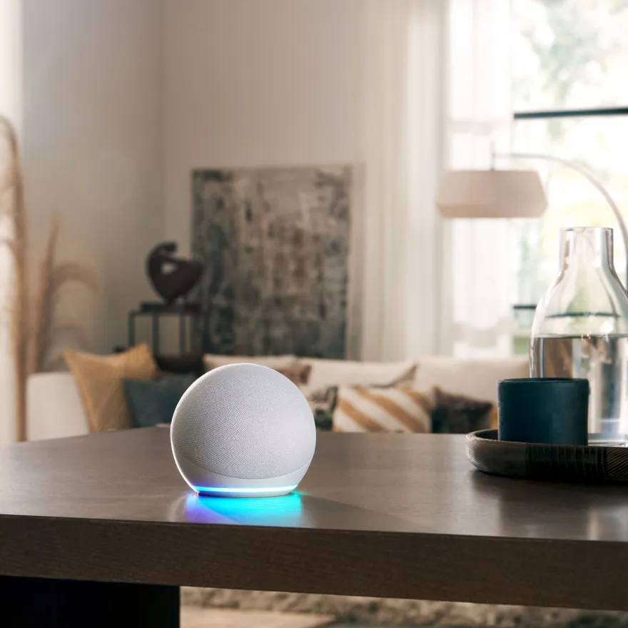 Enceinte intelligente sphérique blanche avec une lumière bleue, posée sur une table en bois dans un salon moderne et lumineux.