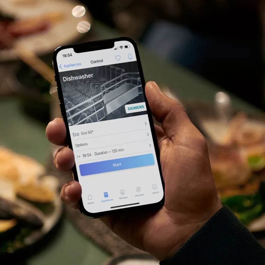 Main tenant un smartphone affichant l'application de contrôle d'un lave‑vaisselle Siemens avec les réglages du cycle Éco et un bouton de démarrage bleu.