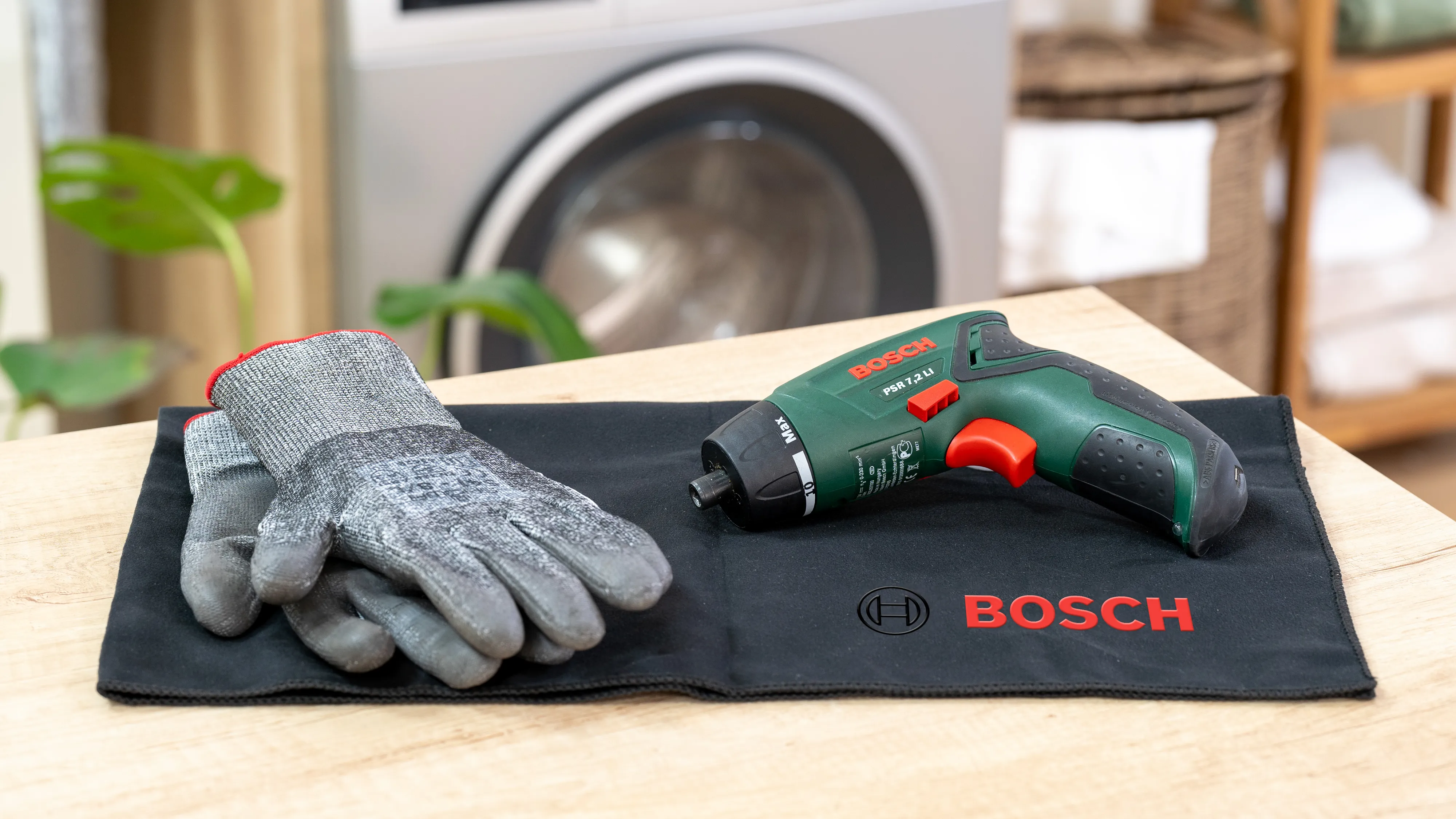 Visseuse Bosch sans fil, gants de travail, et un tissu noir sur une table en bois dans un cadre de maison.