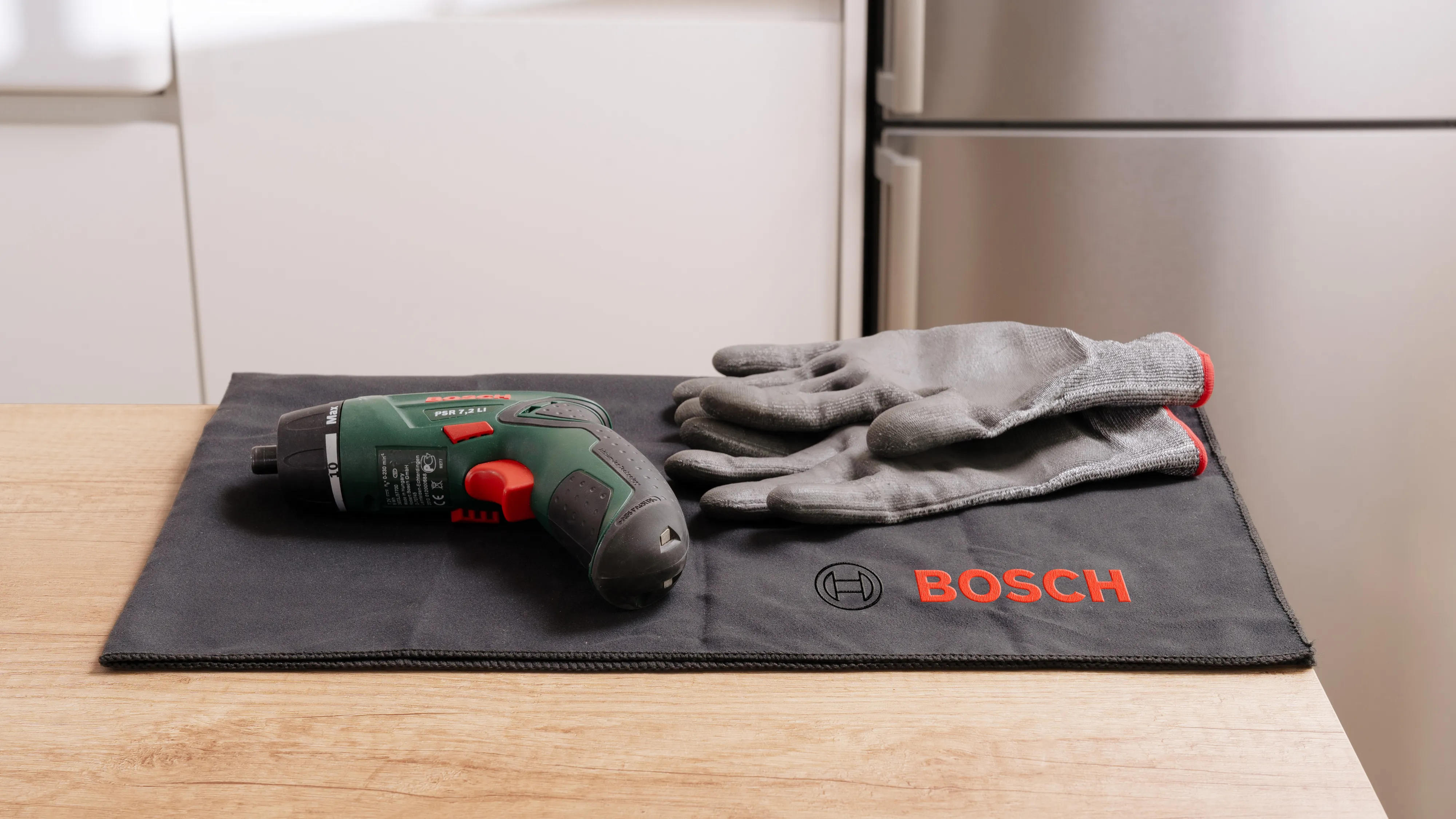 Bosch Bohrer, Handschuhe und Reinigungstuch auf einer Holzoberfläche in der Nähe von Küchengeräten.