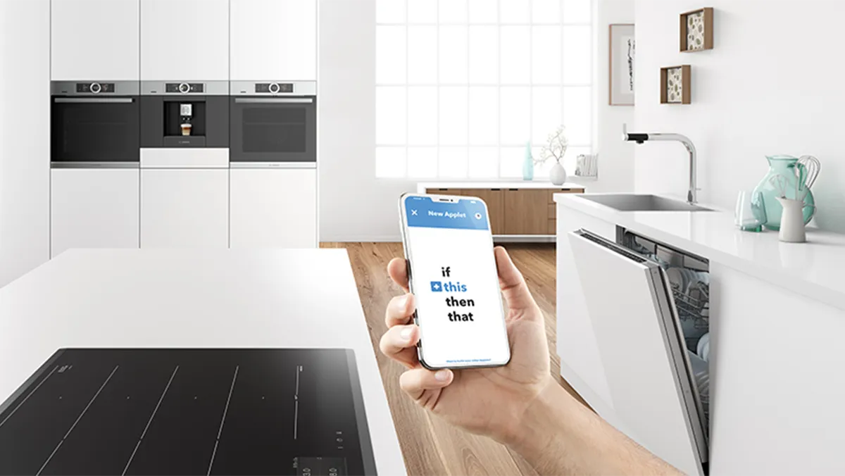 Eine Hand hält ein Smartphone mit der IFTTT-App zur Automatisierung von Bosch Hausgeräten in einer modernen weißen Küche.