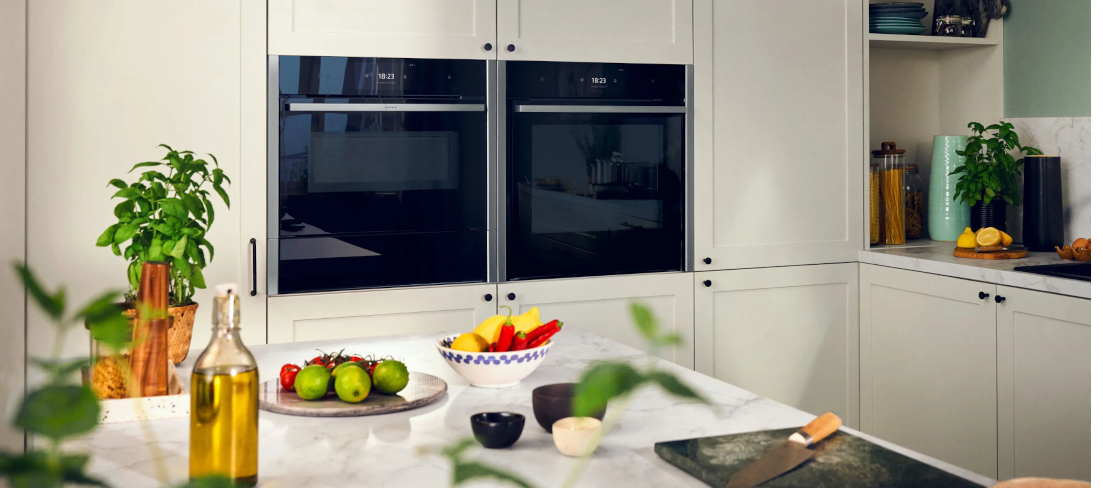 Une personne en chemise blanche tient un verre de liquide vert dans une cuisine moderne, drapée d'un torchon à motif floral.