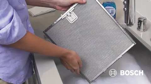 Person cleaning a Bosch range hood filter in a sink with soap and water.