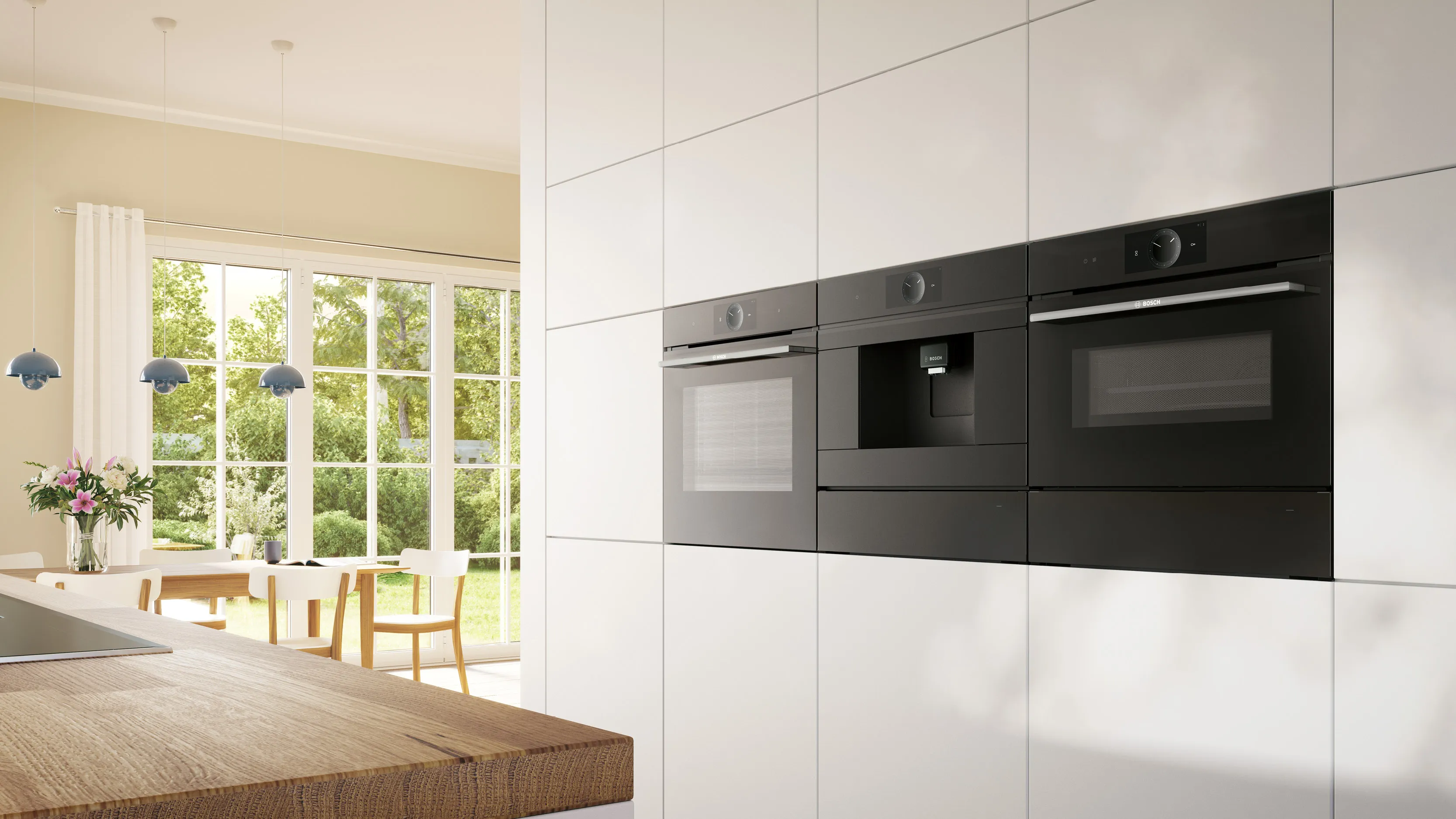A bright, modern kitchen with integrated black Bosch appliances (oven, coffee machine, microwave) set into white cabinetry, with a dining area and large windows overlooking a green garden.