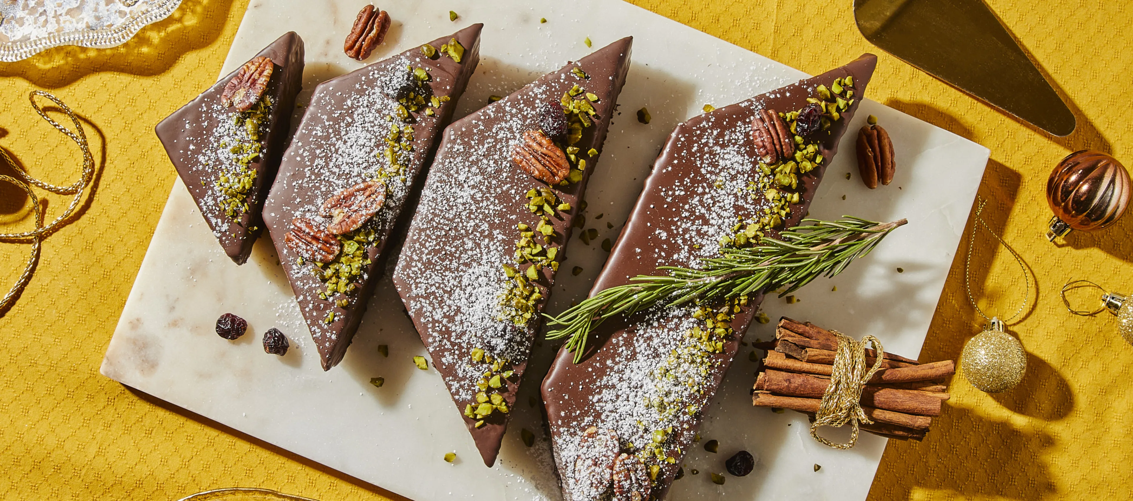 Chocolate gingerbread triangles with powdered sugar, pistachios, and cinnamon sticks on a yellow background.
