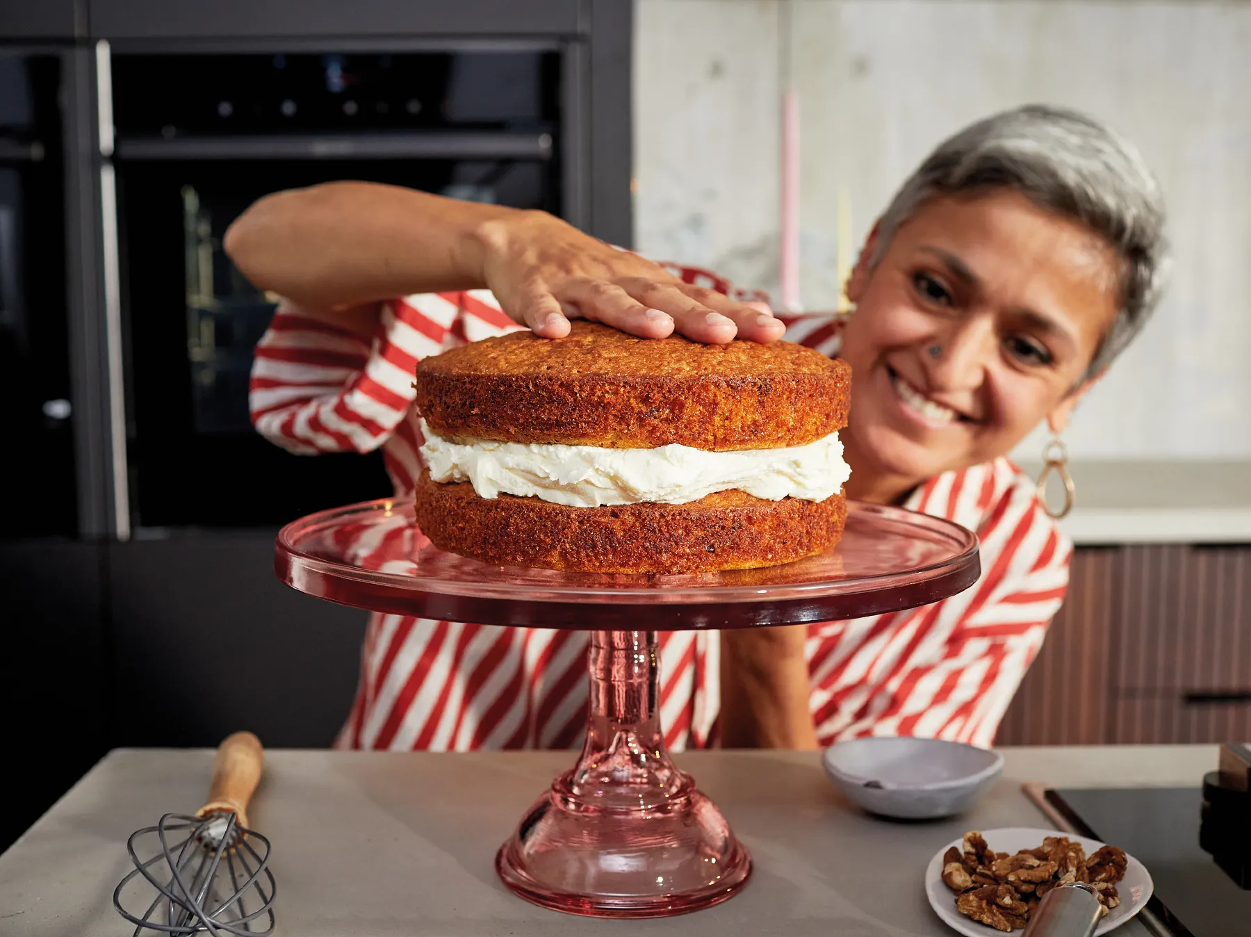 A smiling woman with short grey hair, wearing a red and white striped shirt, is assembling a two-layer cake in a modern kitchen. She gently places the top, golden-brown layer onto a bottom layer generously frosted with white cream. The cake rests on a pink glass cake stand. A whisk and a plate of walnuts are visible on the light countertop in the foreground. Dark built-in ovens are in the blurred background, along with colorful candles.