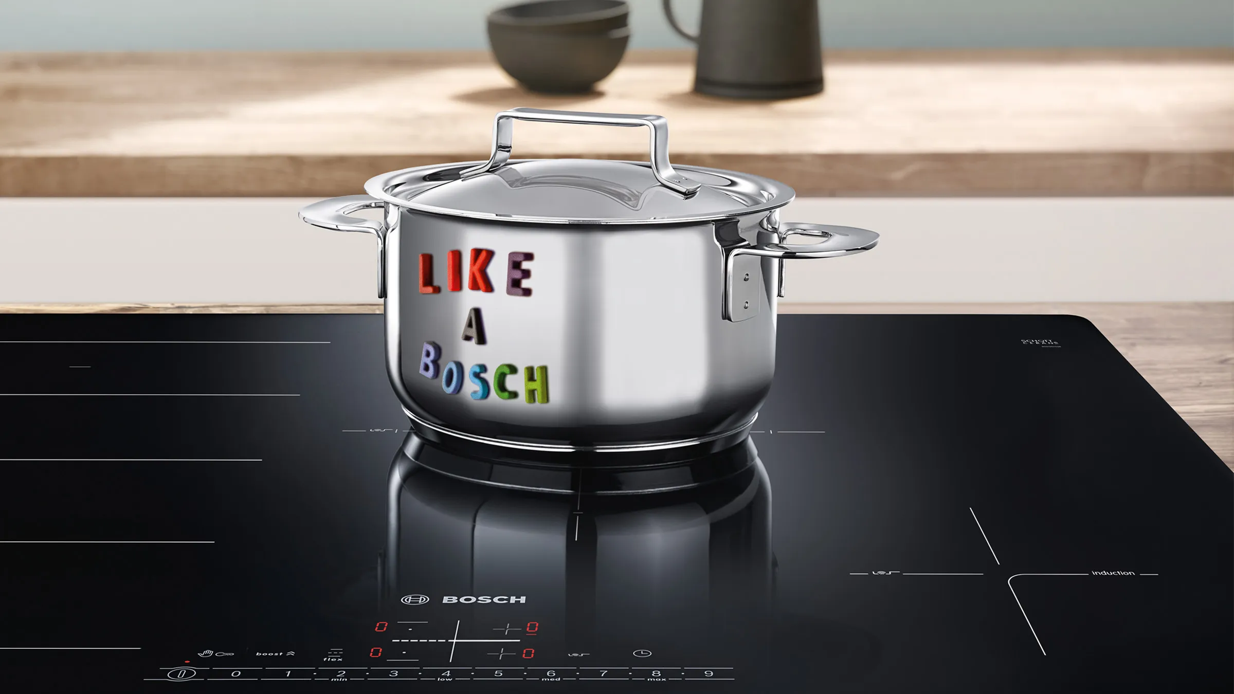 A stainless steel pot with letter magnets spelling "like a bosch" sits on an induction cooktop in a bright kitchen.