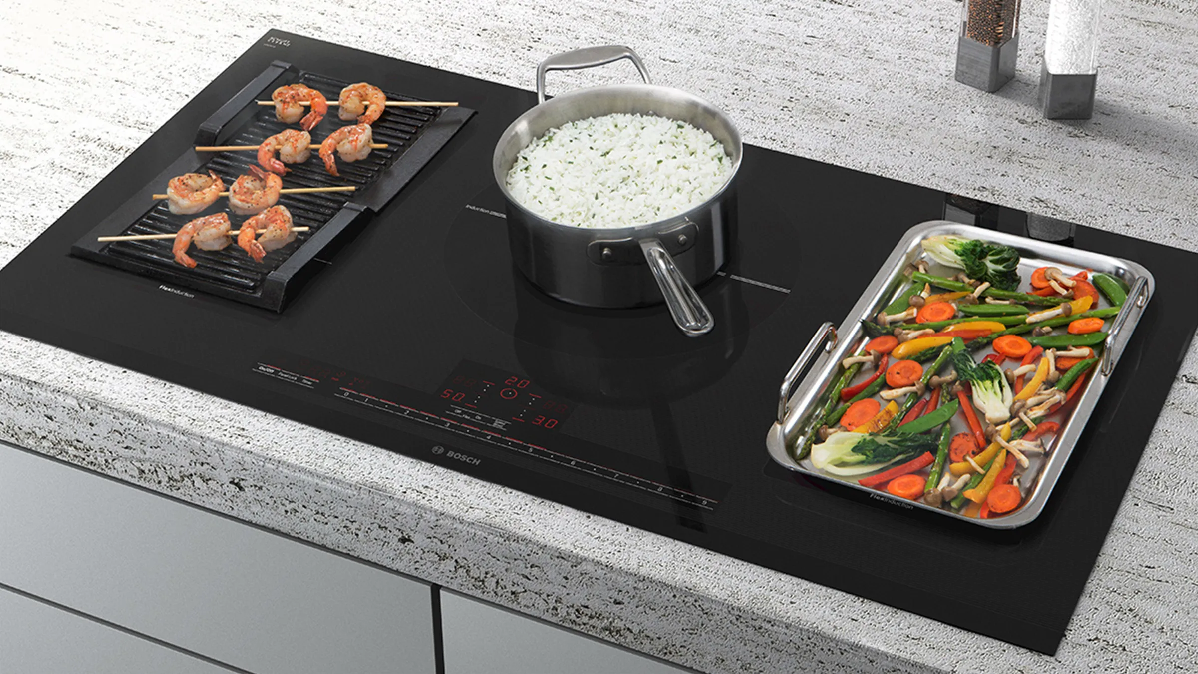 A kitchen countertop with an integrated induction cooktop featuring teppanyaki with vegetables, a grill plate with prawn skewers, and a pot of rice in the center.