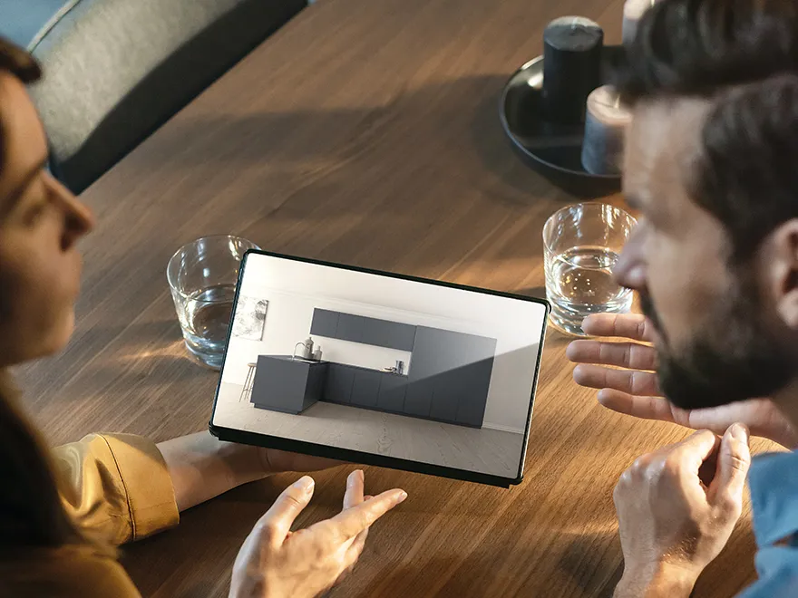 Osoba ukazuje design kuchyně na tabletu a diskutuje o něm s jinou osobou, sedící u dřevěného stolu se sklenicemi vody.