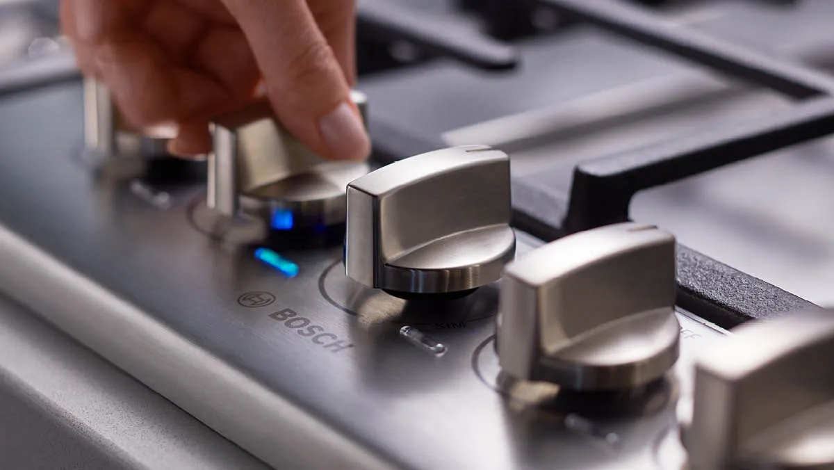 Close-up of a sleek stovetop control panel with three silver knobs and digital flame level indicator.