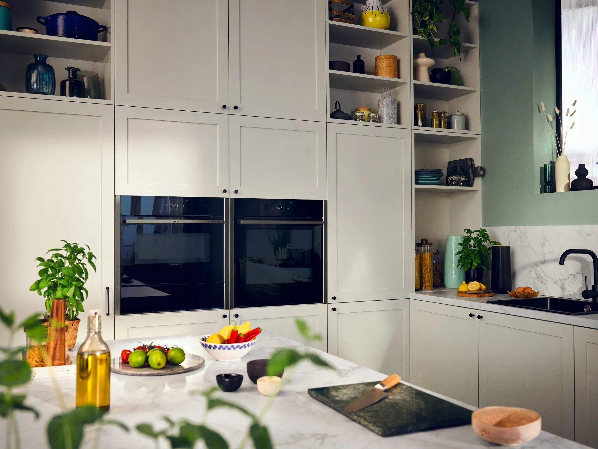 Modern kitchen with white cabinets, built-in ovens, shelves, and fresh produce on the counter.