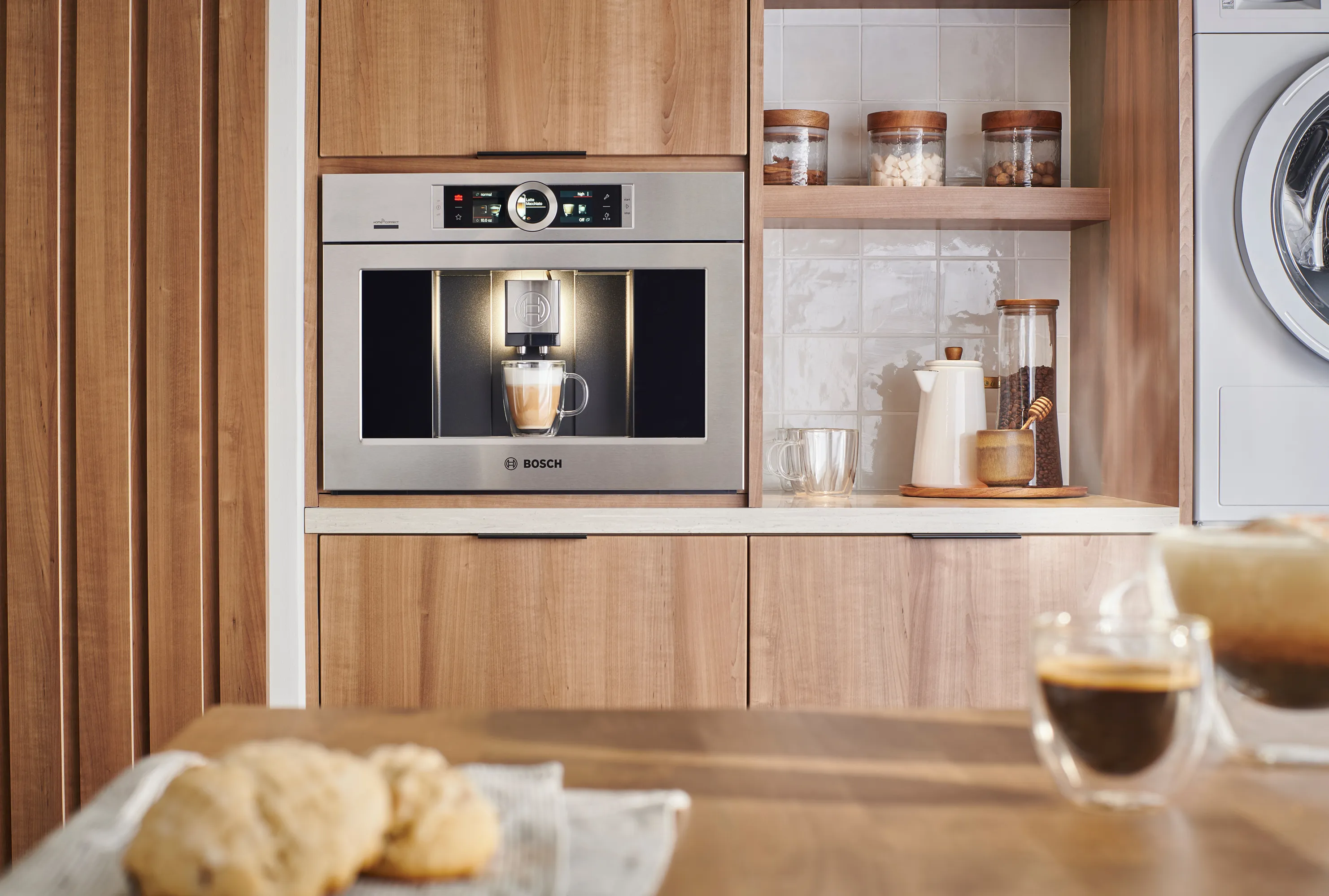 A modern Bosch coffee machine in a wooden kitchen, preparing coffee with cookies on a table in the foreground.